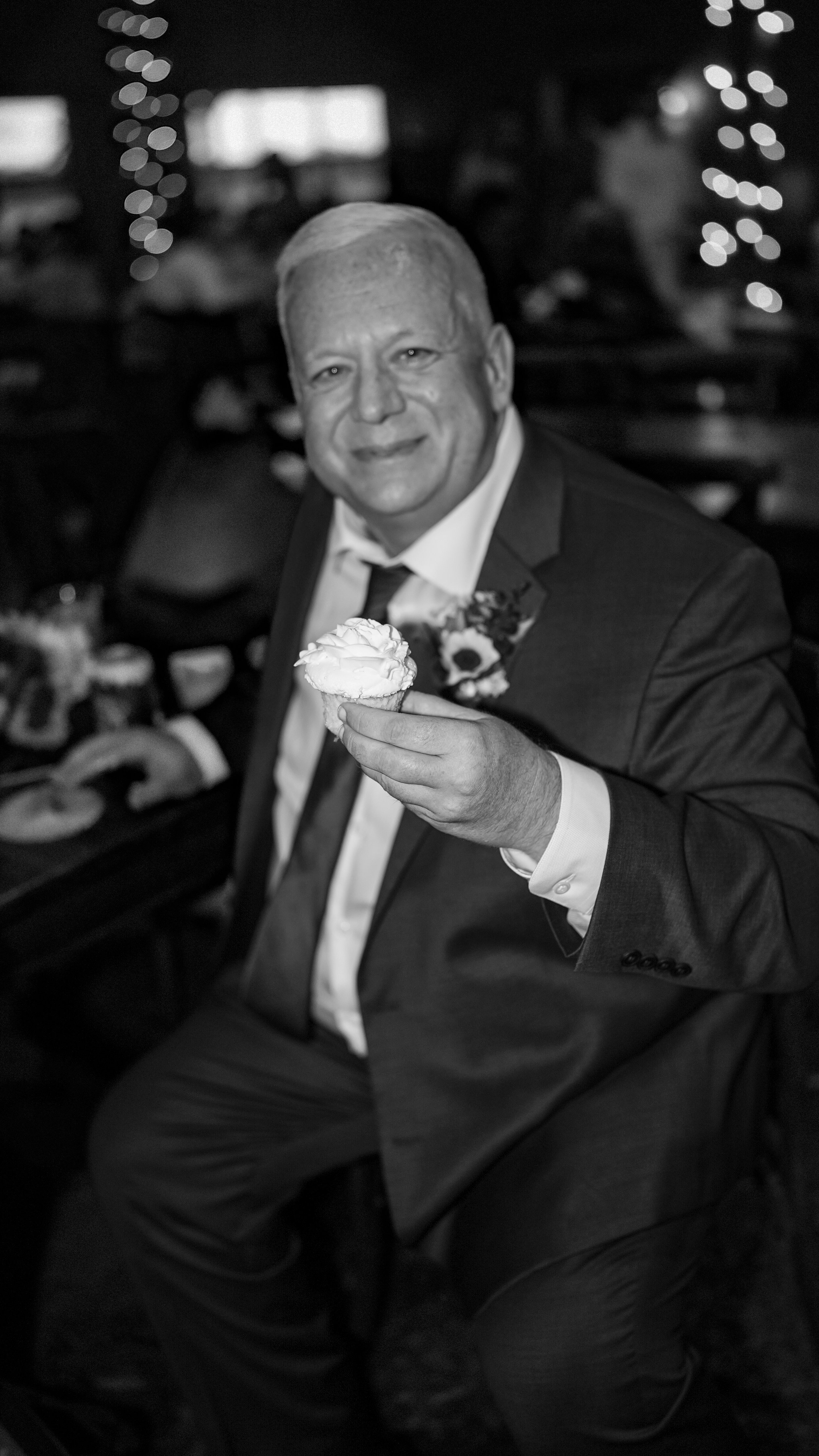 A smiling man in a tuxedo holding a cupcake at a celebration or party.