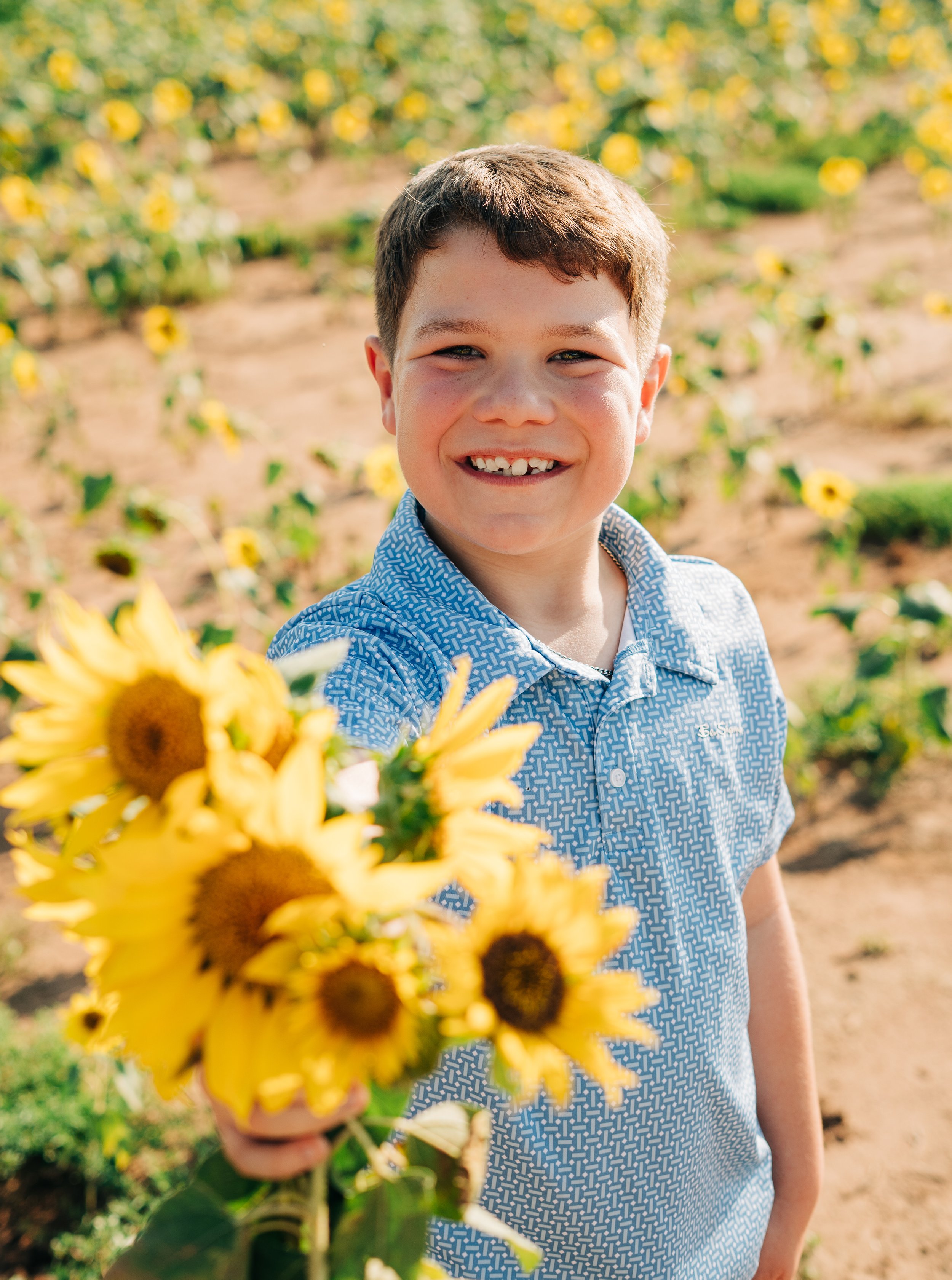 Smiling boy with missing tooth holding a bouquet of yellow flowers in a sunflower field. Eastern Shore photography. Family photography. Graduation Photography. Couples Photography in Fairhope, Daphne, and Spanish Fort.