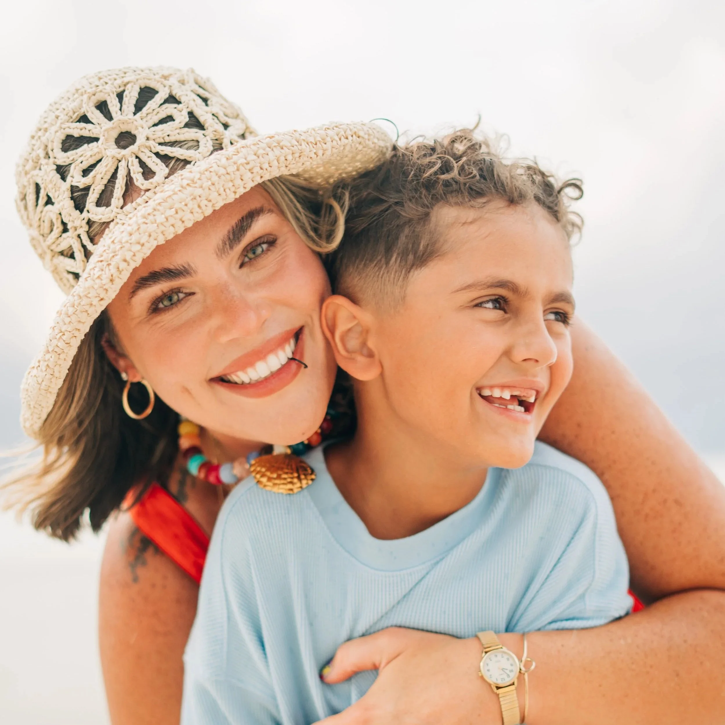 Close-up of a woman smiling, wearing a large straw sunhat with crochet details and a multicolored beaded necklace, with a young person next to her. Family Photography in Baldwin County.