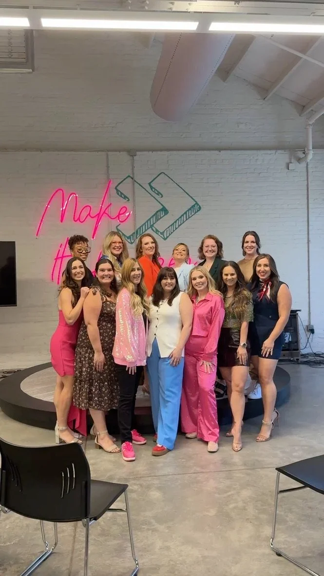 Group of women posing together on a stage with a white brick wall background, neon pink and green sign