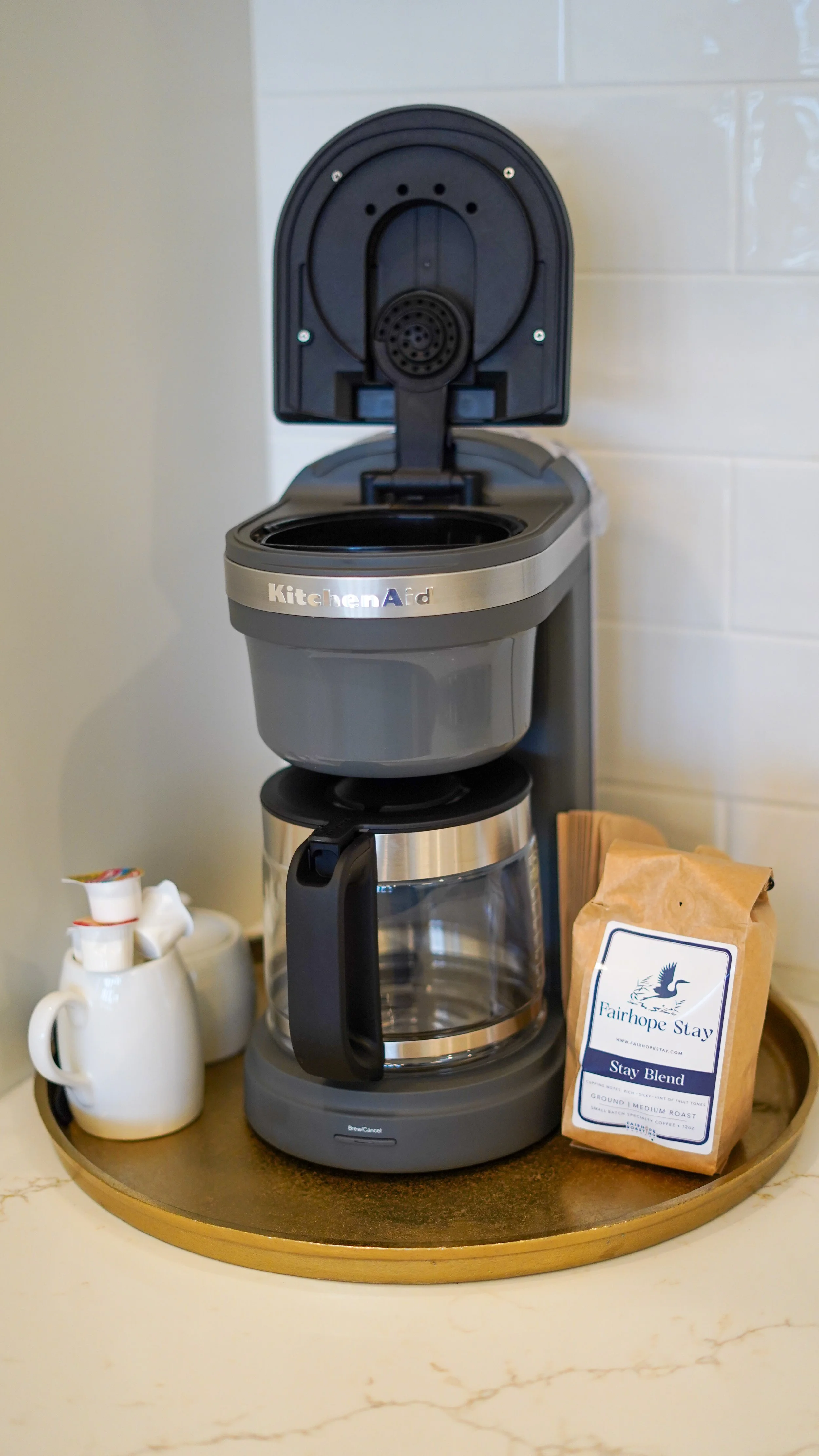 A coffee maker with an open top, a glass carafe, and a bag of Fairhope Stay coffee on a round wooden tray, with creamer cups nearby.