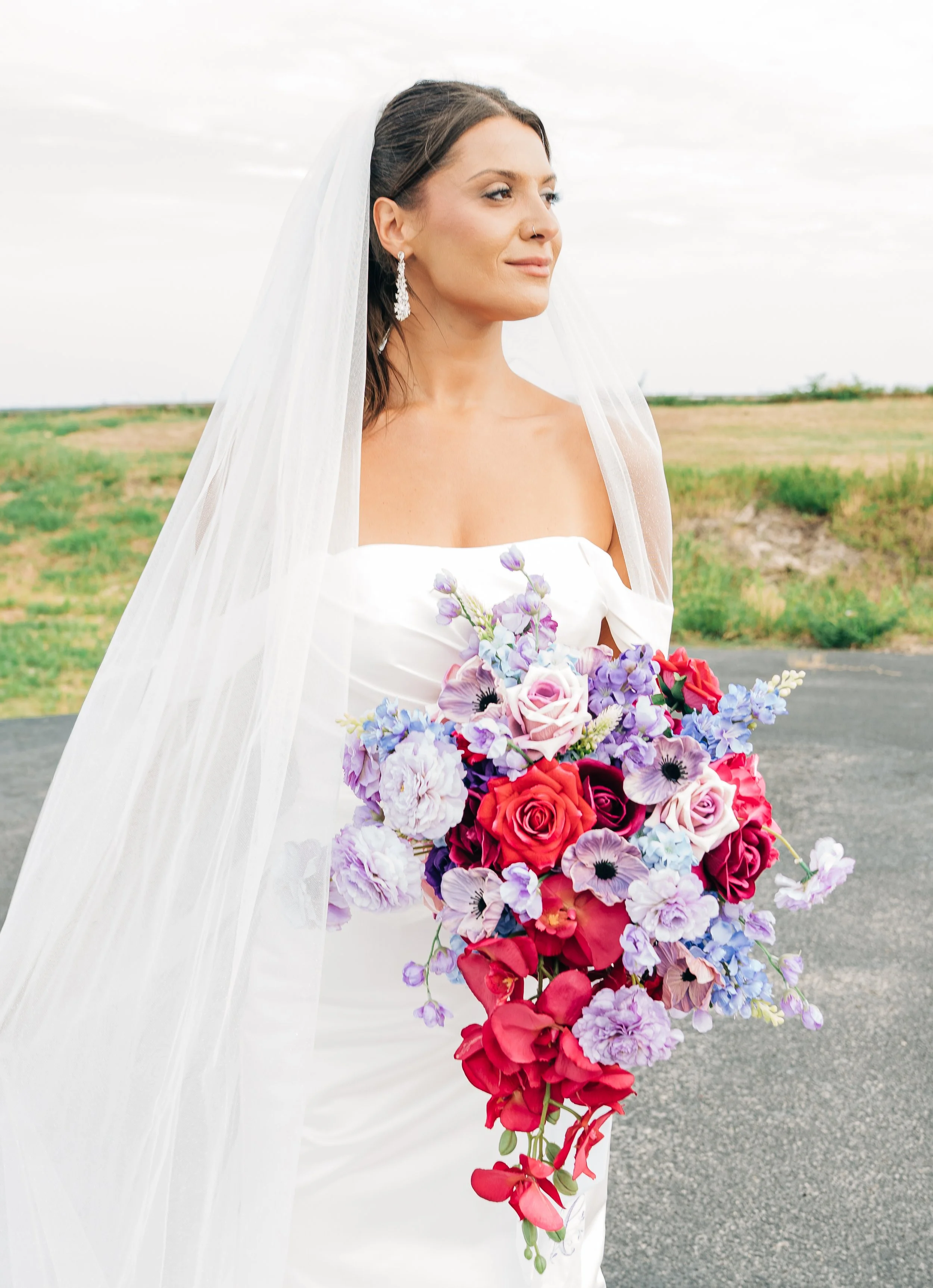 A bride in a white wedding dress holding a large bouquet of colorful flowers, standing outdoors with a cloudy sky and open land in the background. Wedding Photography in Gulfport, Mississippi.