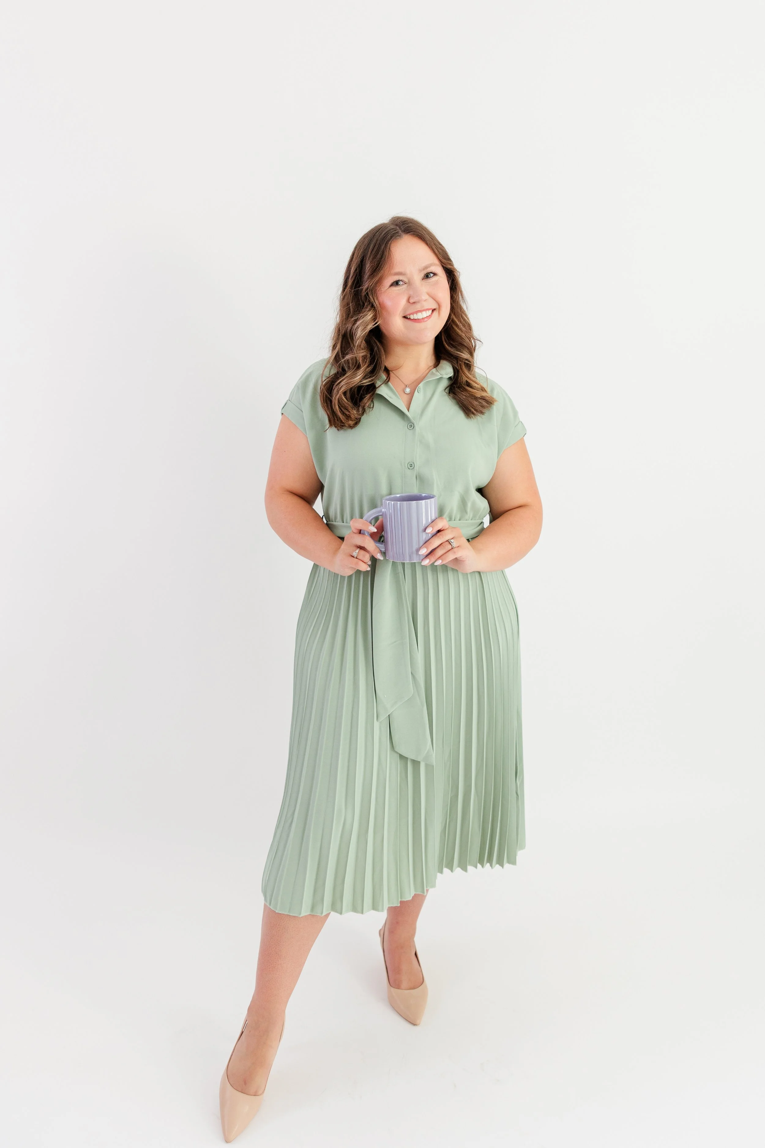 Woman in a light green blouse and pleated skirt holding a purple mug, standing against a white background.