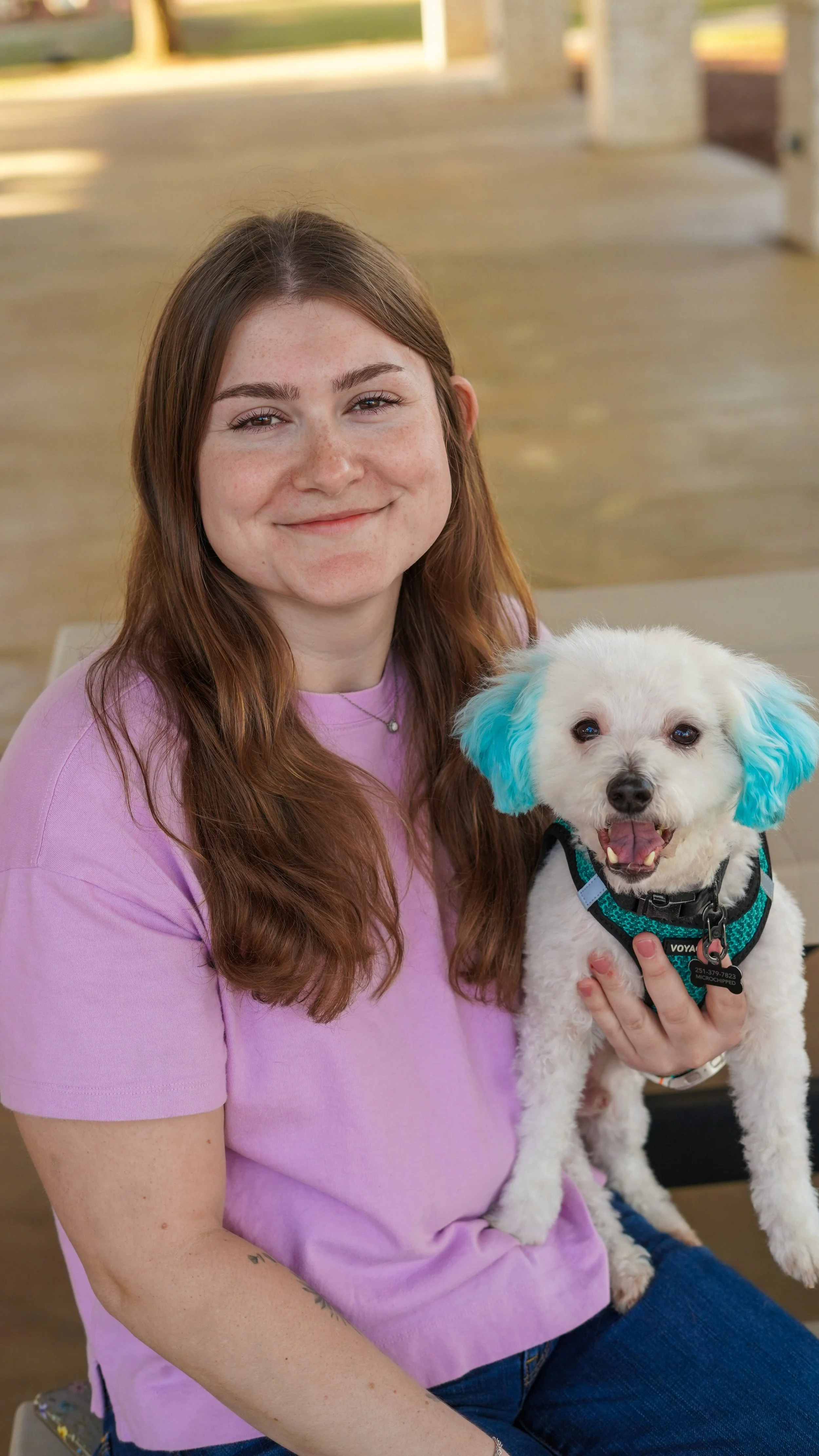 A young woman with long brown hair and a purple shirt holding a small white dog with blue ears inside a covered outdoor area.