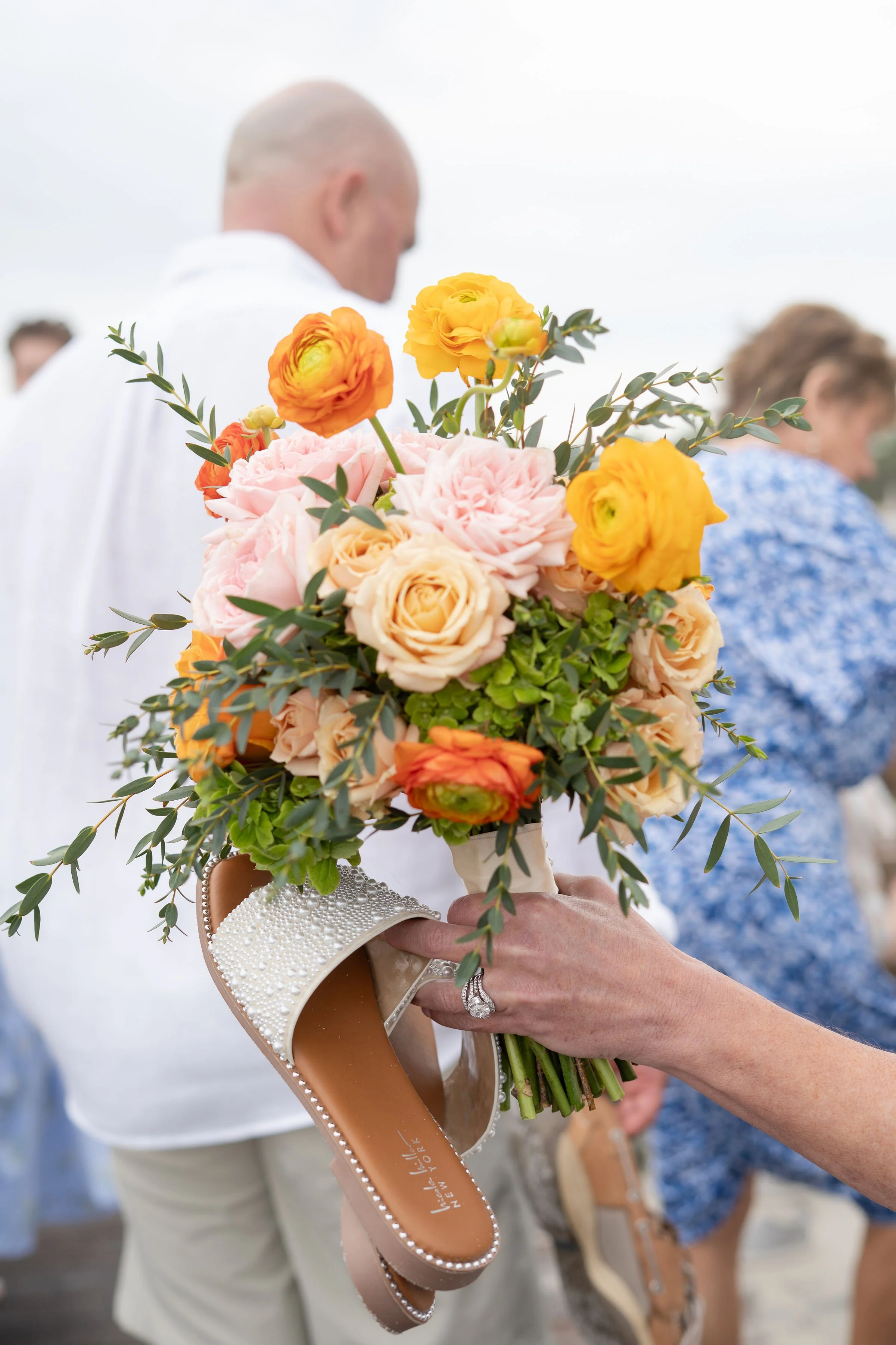A bouquet of pink, yellow, and orange flowers with green foliage being held by a person wearing a ring, with a blurred man and woman in the background at an outdoor event.
