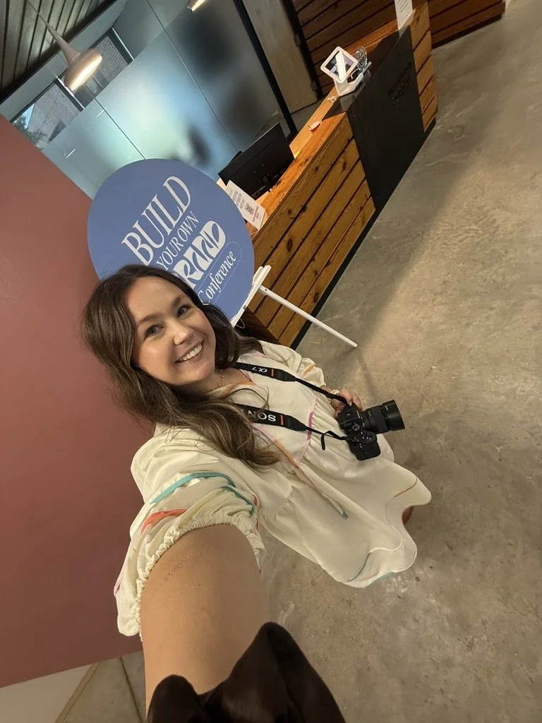 A woman taking a selfie in an indoor setting, smiling, with a camera around her neck, standing in front of a wooden table with a sign that says 'Build Your Own AR/VR Conference'.