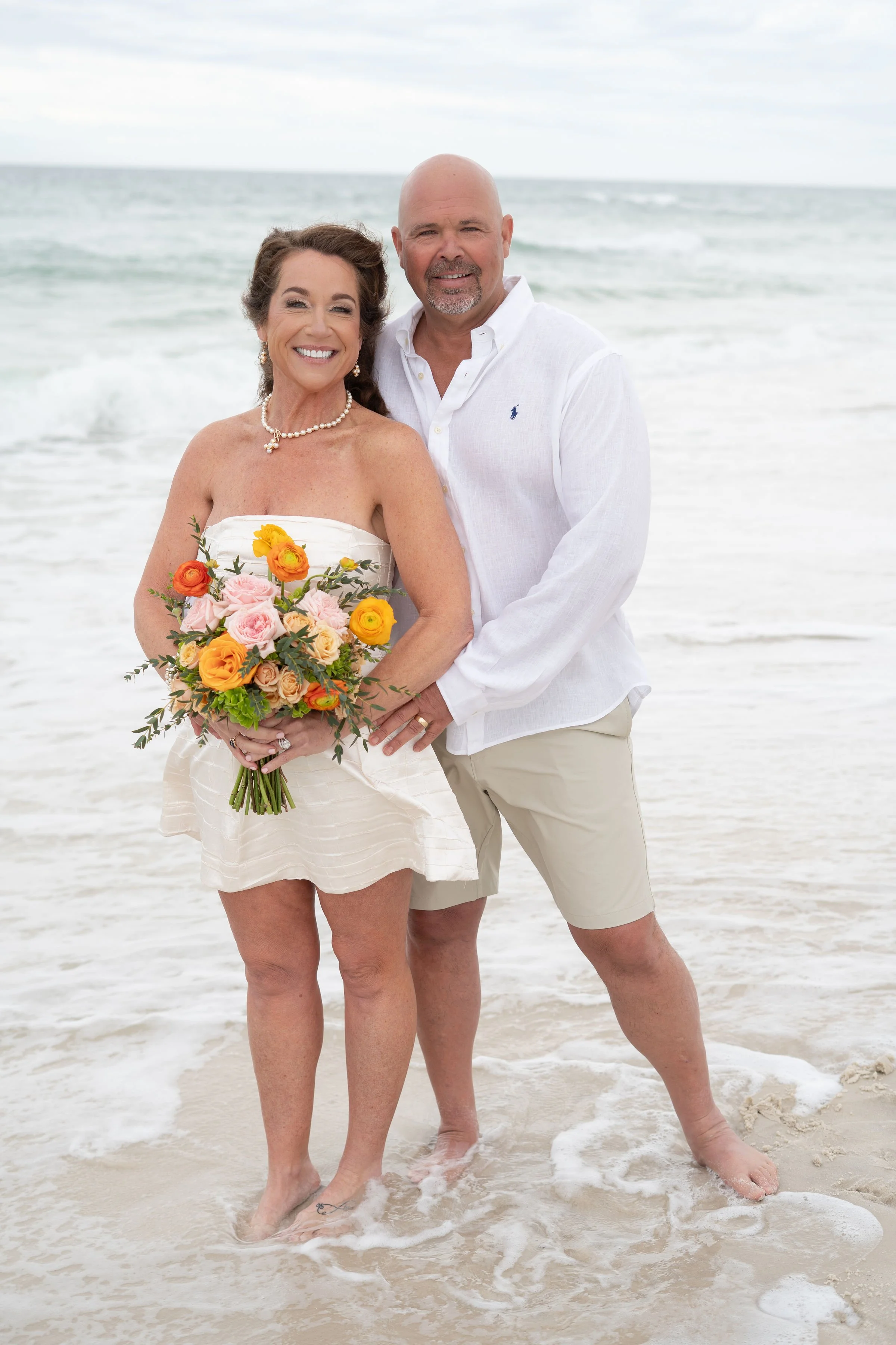 A smiling woman holding a colorful bouquet standing next to a smiling man on a beach with ocean waves in the background.