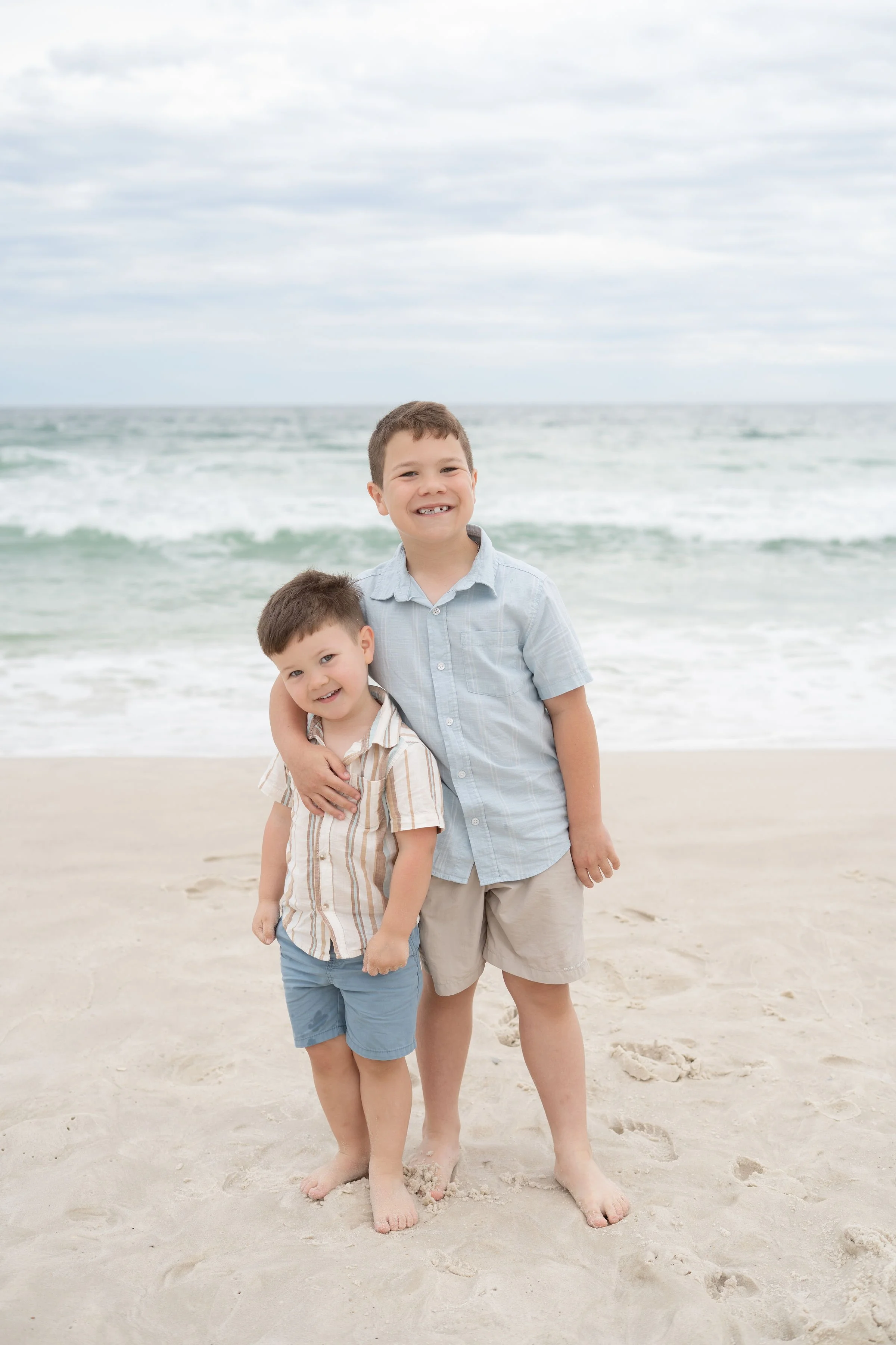 Two young boys standing on a sandy beach, smiling with arms around each other, ocean waves and cloudy sky in the background.