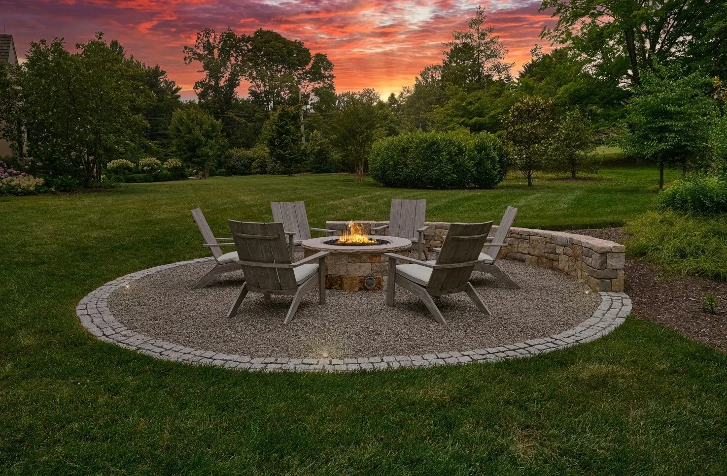 A backyard scene at sunset with a stone fire pit surrounded by six wooden chairs on a gravel circle, with lush green grass and trees in the background.