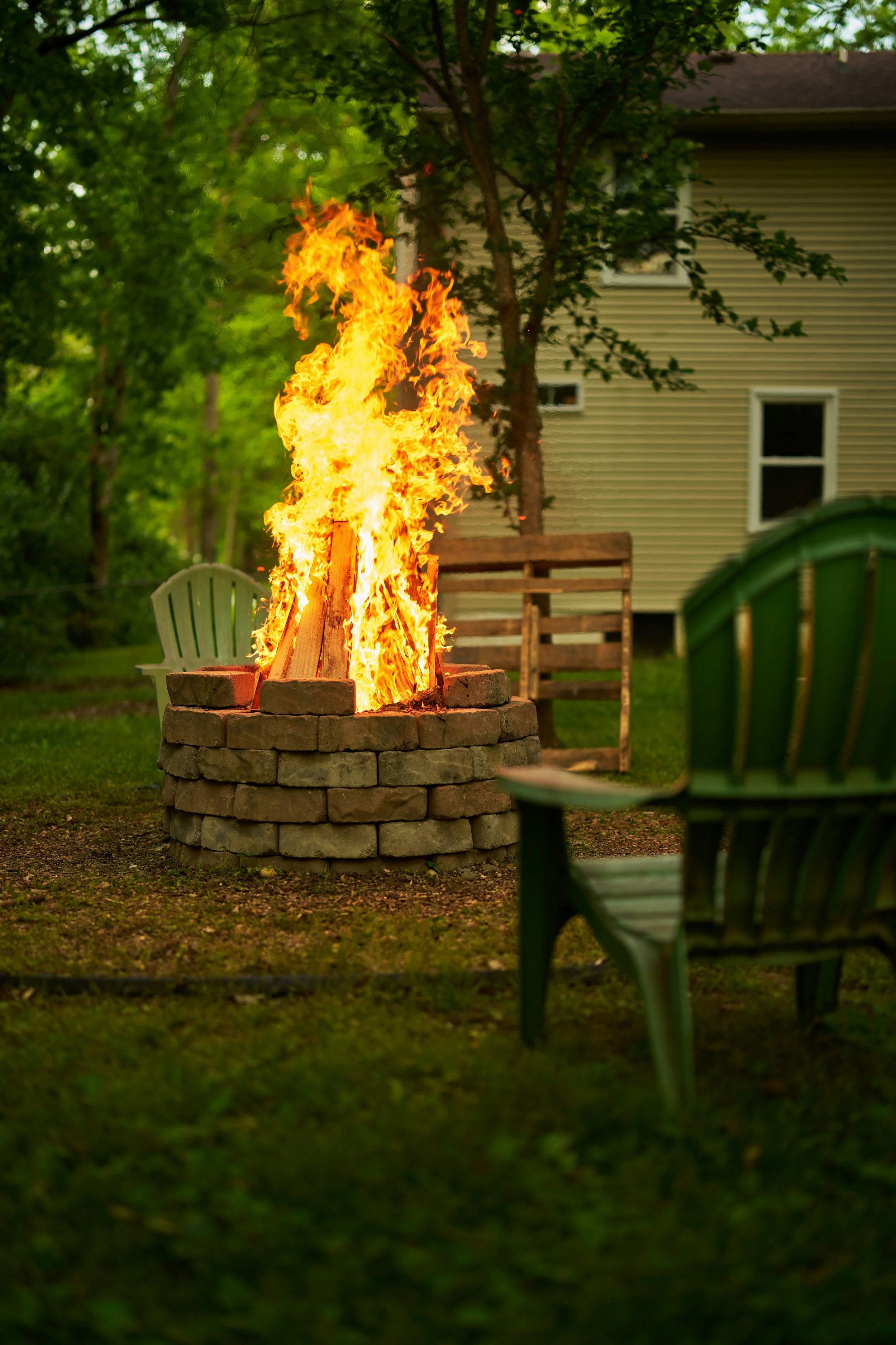 Outdoor backyard scene with a controlled campfire in a stone firepit, surrounded by green chairs, trees, and a house in the background.