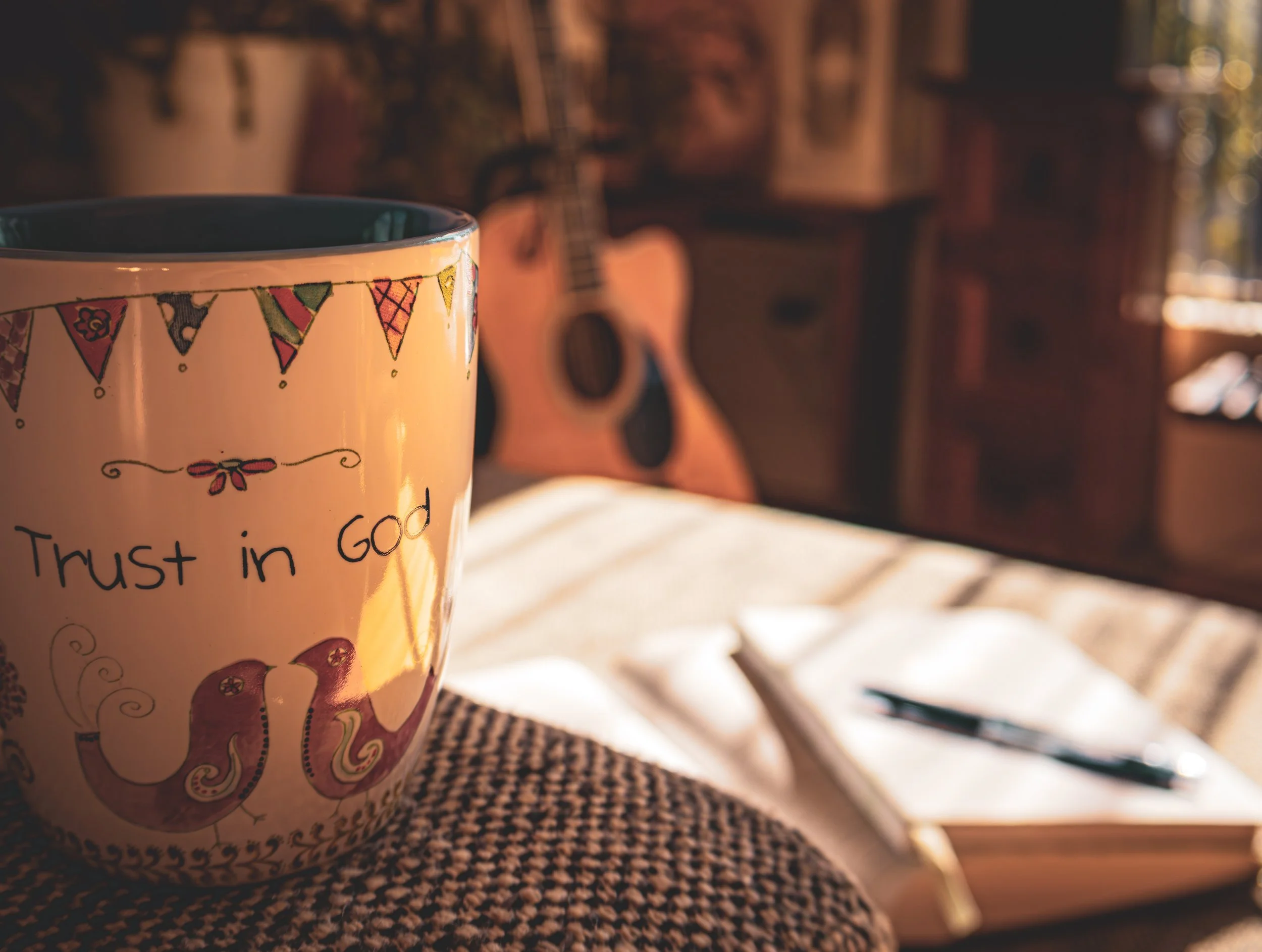 A mug with "trust in God", an open notebook blurred in the background and a guitar on a stand in the corner, with natural sun light shining through the window