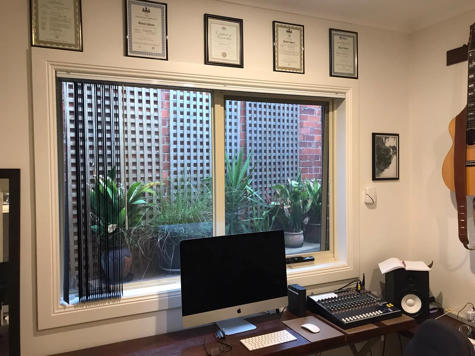 Home recording studio with iMac, audio mixing console, studio monitor speaker, framed certificates, and a plant-filled courtyard visible through the window