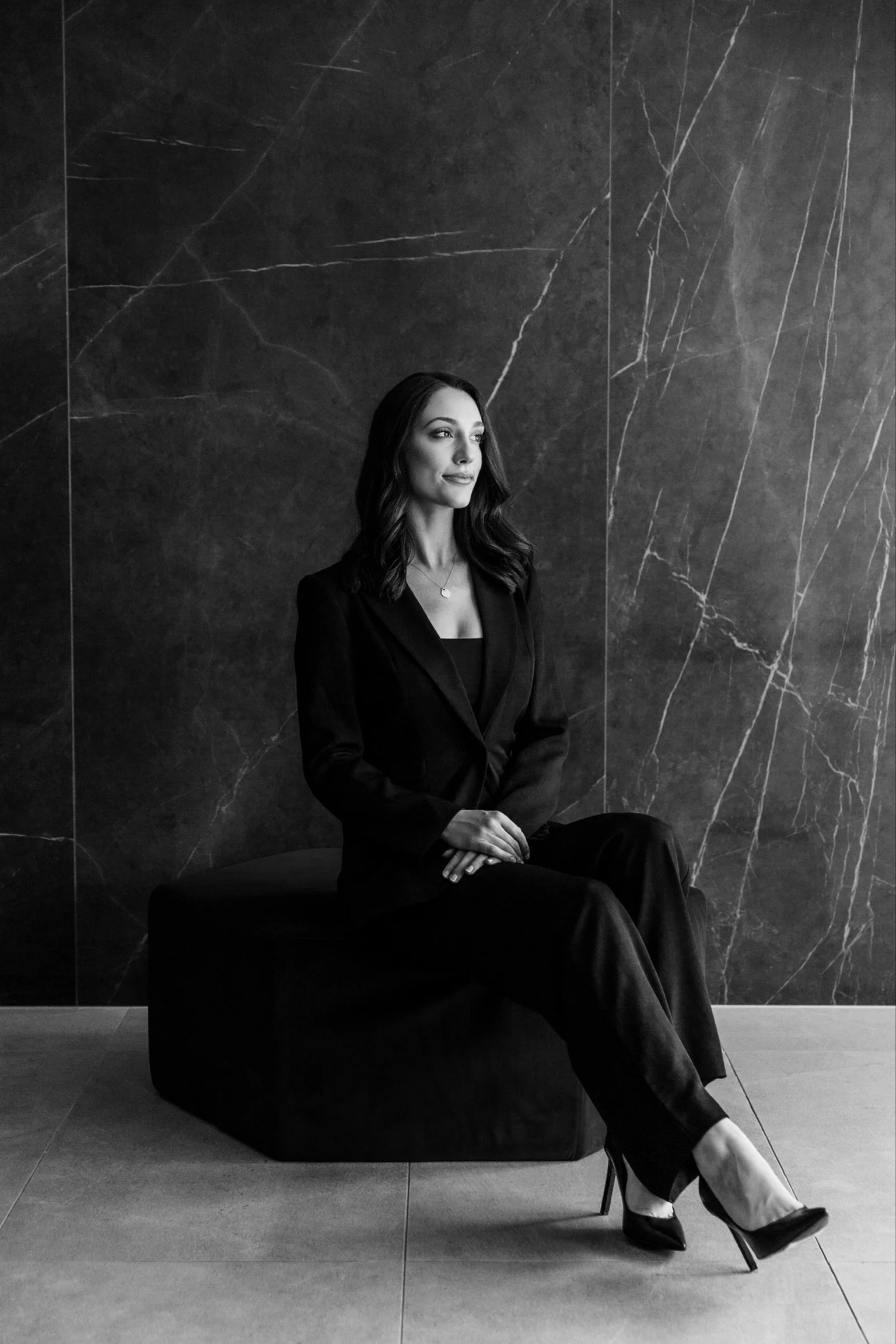 A woman with dark hair, dressed in a black suit and high heels, sitting on a black bench against a dark marble wall.