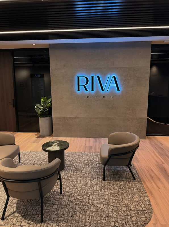 Lobby area with a large sign reading 'RIVA OFFICES' on a wall, surrounded by beige and black furniture, a potted plant, and a patterned rug.
