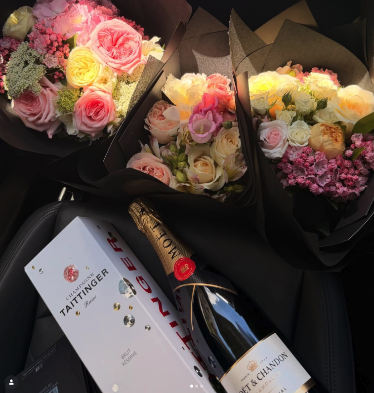 Three bouquets of pink, white, and yellow roses with greenery, a box of Taittinger champagne, a bottle of Veuve Clicquot champagne, all placed on a black surface inside a car.