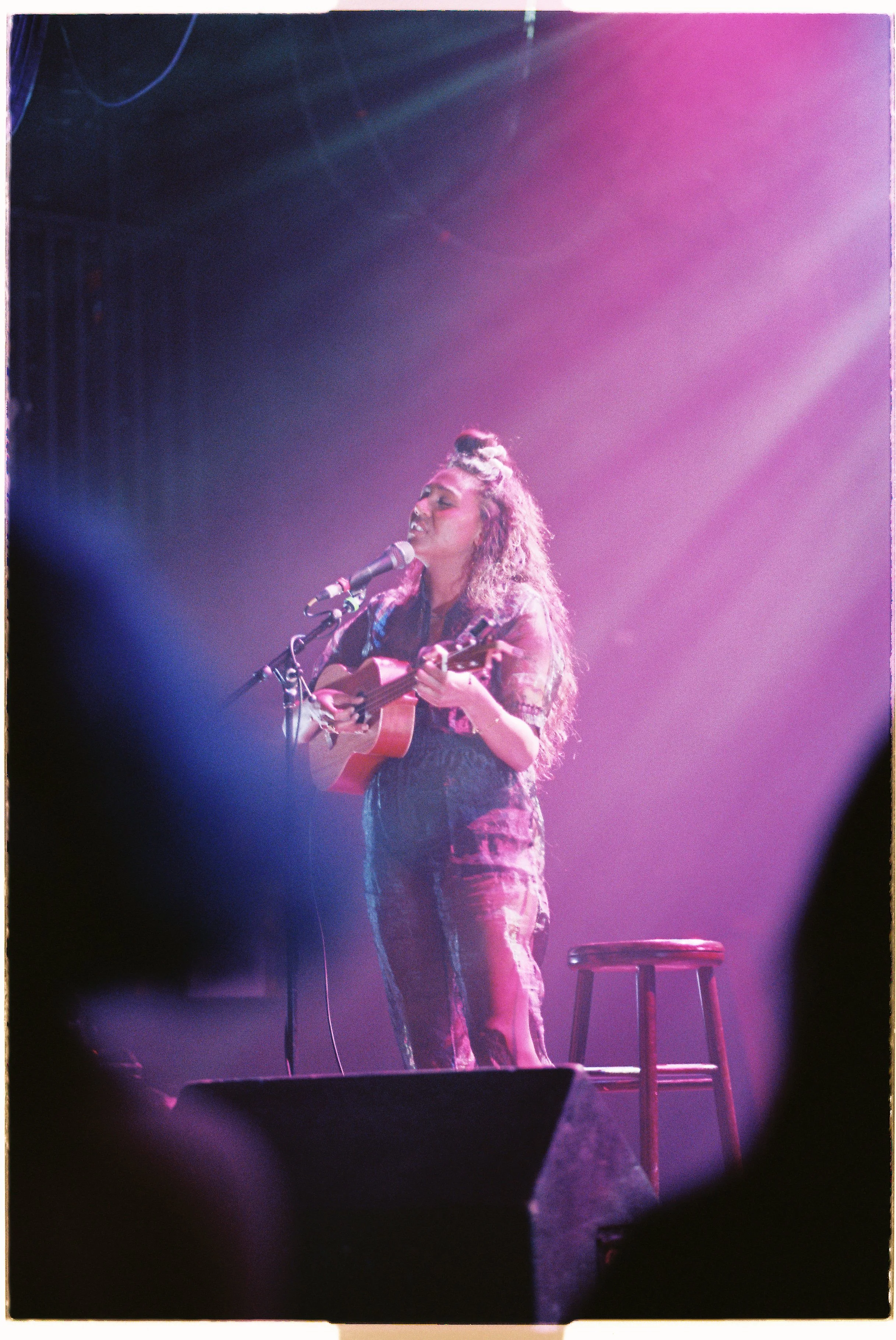 Desirée Dawson with curly hair and a top knot hairstyle is performing solo on stage with a small guitar and singing into a microphone, illuminated by pink and purple stage lights, with a stool and speakers visible in the background.