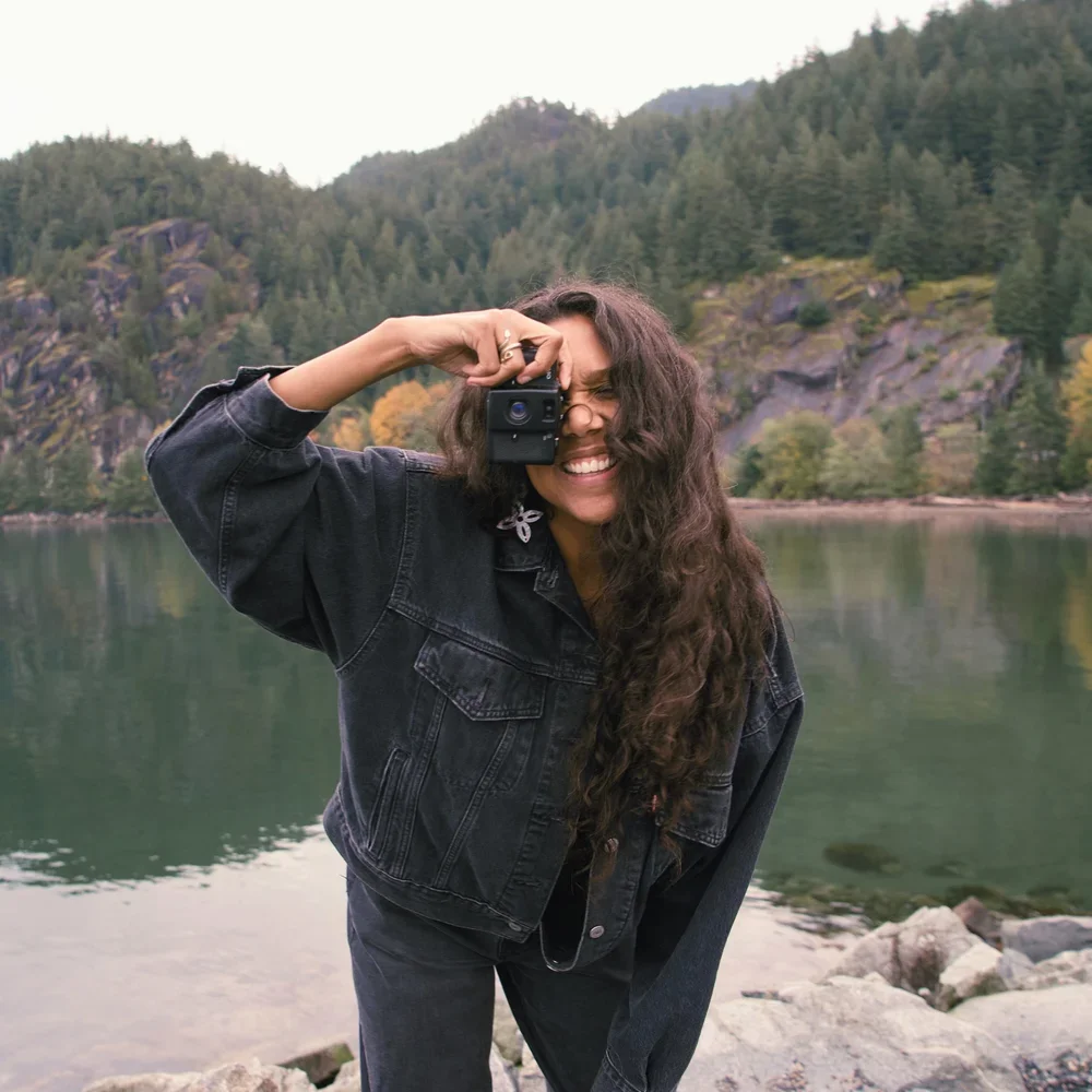 Desirée Dawson with long curly hair taking a photograph with a camera by a lake surrounded by lush green mountains.