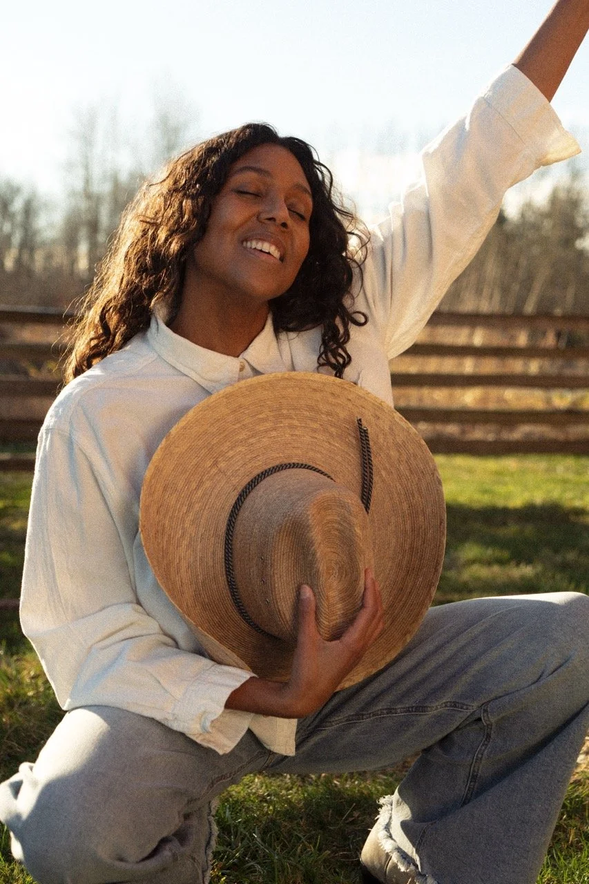 Desirée Dawson in a beige shirt and jeans sitting outdoors on grass, holding a wide-brimmed straw hat with her eyes closed, smiling, and enjoying the sunny weather.