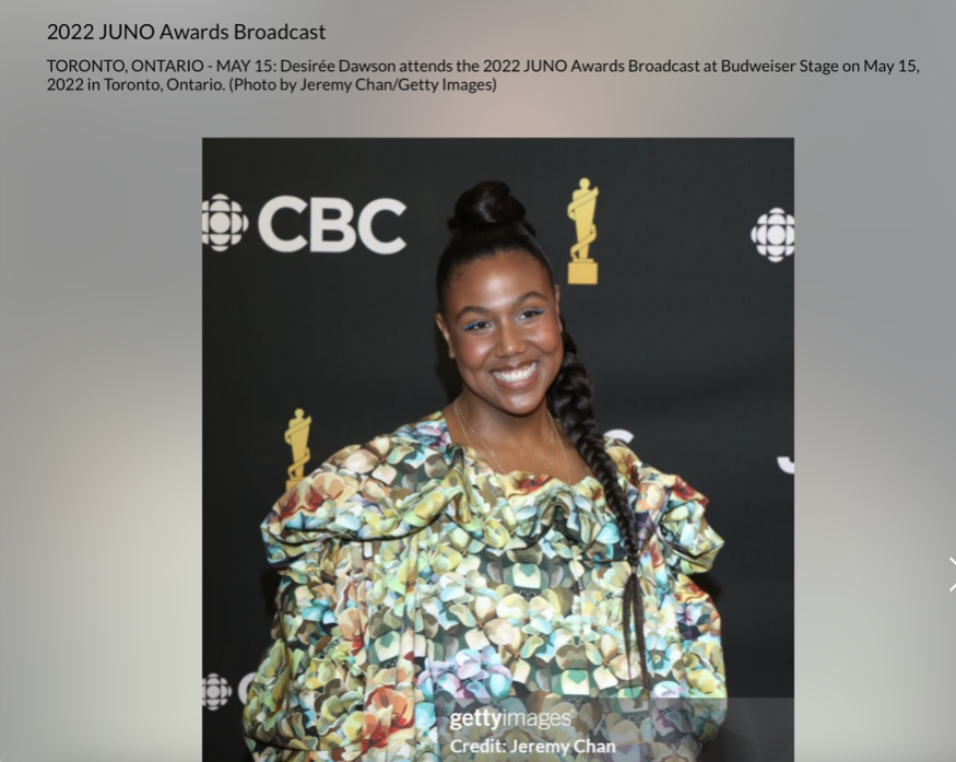 Desirée Dawson smiling at the JUNO Awards, wearing a colorful, floral-patterned dress, with a high bun hairstyle, standing in front of a black background with logos for CBC and an award statue.