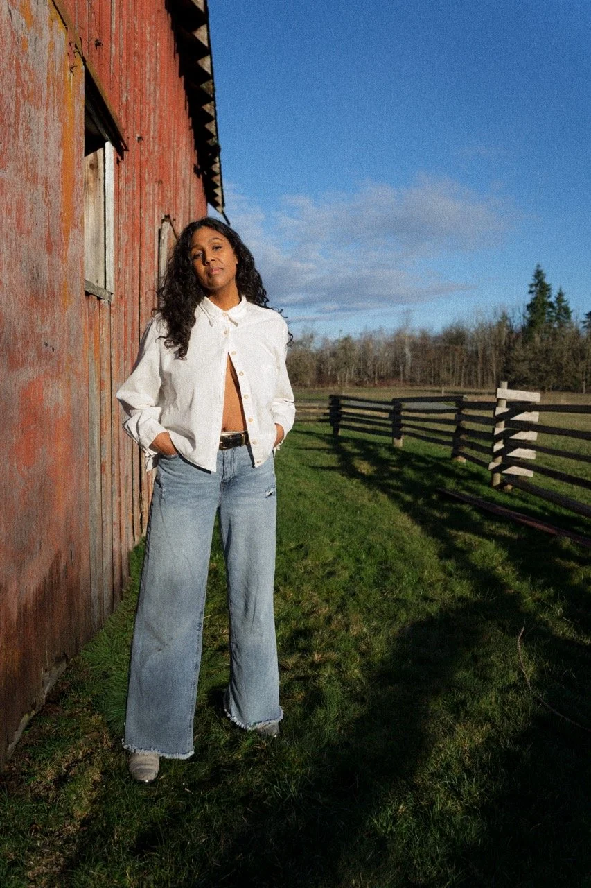 Desirée Dawson with long curly hair in a white shirt and wide-leg jeans standing outdoors beside a weathered wooden barn, under a bright blue sky with some clouds, and a wooden fence in the background.