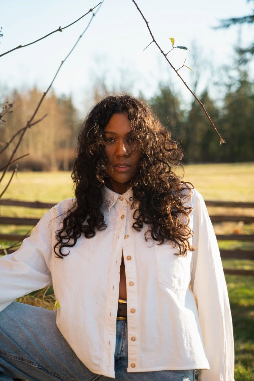 Desirée Dawson with curly hair sitting outdoors next to a wooden fence, wearing a white button-up shirt and jeans, with trees and a fence in the background.
