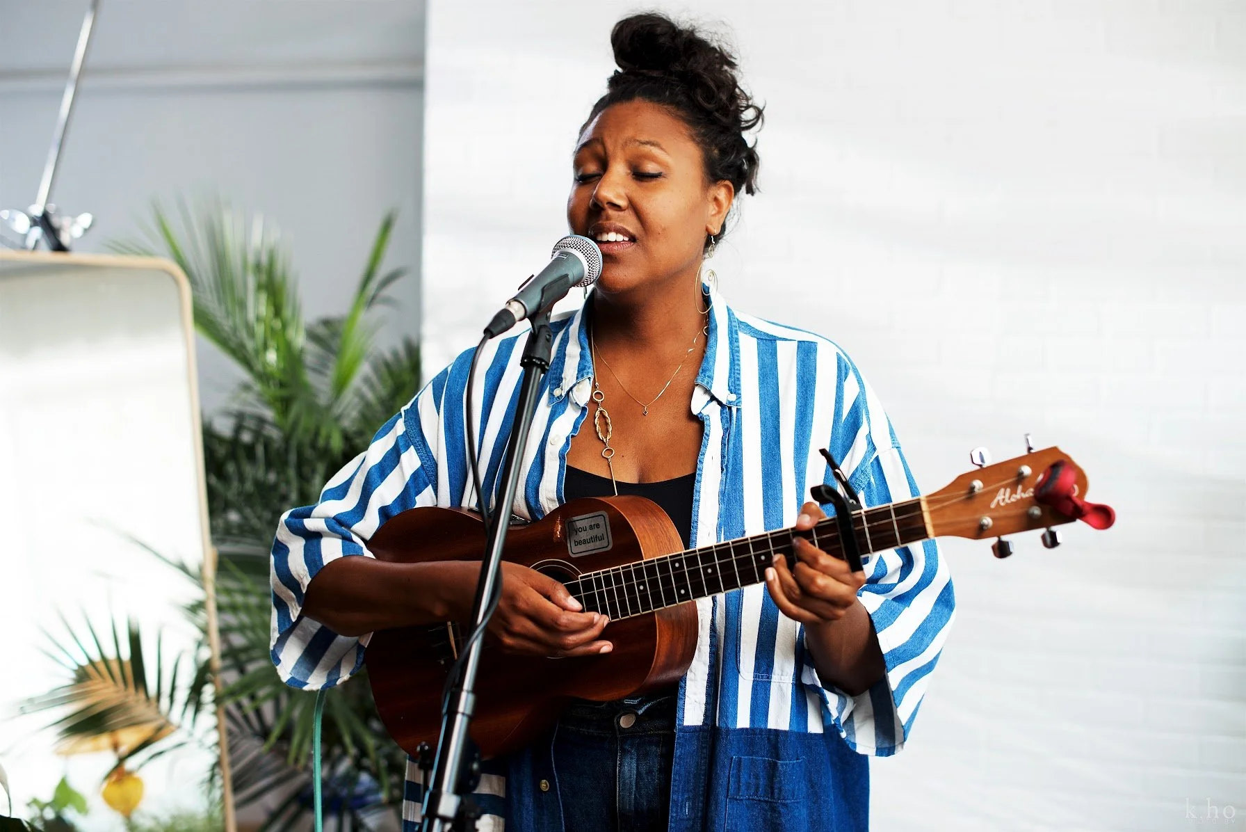 Desirée Dawson singing and playing baritone ukulele in front of a microphone, wearing a blue and white striped shirt.
