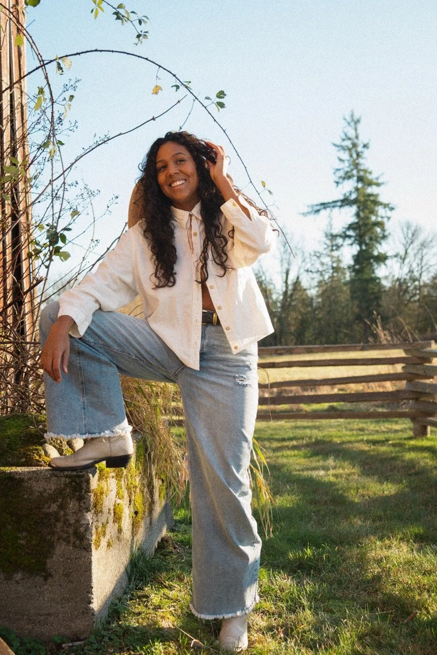 Desirée Dawson with curly hair in white shirt and ripped jeans poses outdoors, leaning on a moss-covered concrete block with one foot on it, smiling, with a wooden fence and trees in the background during daylight.