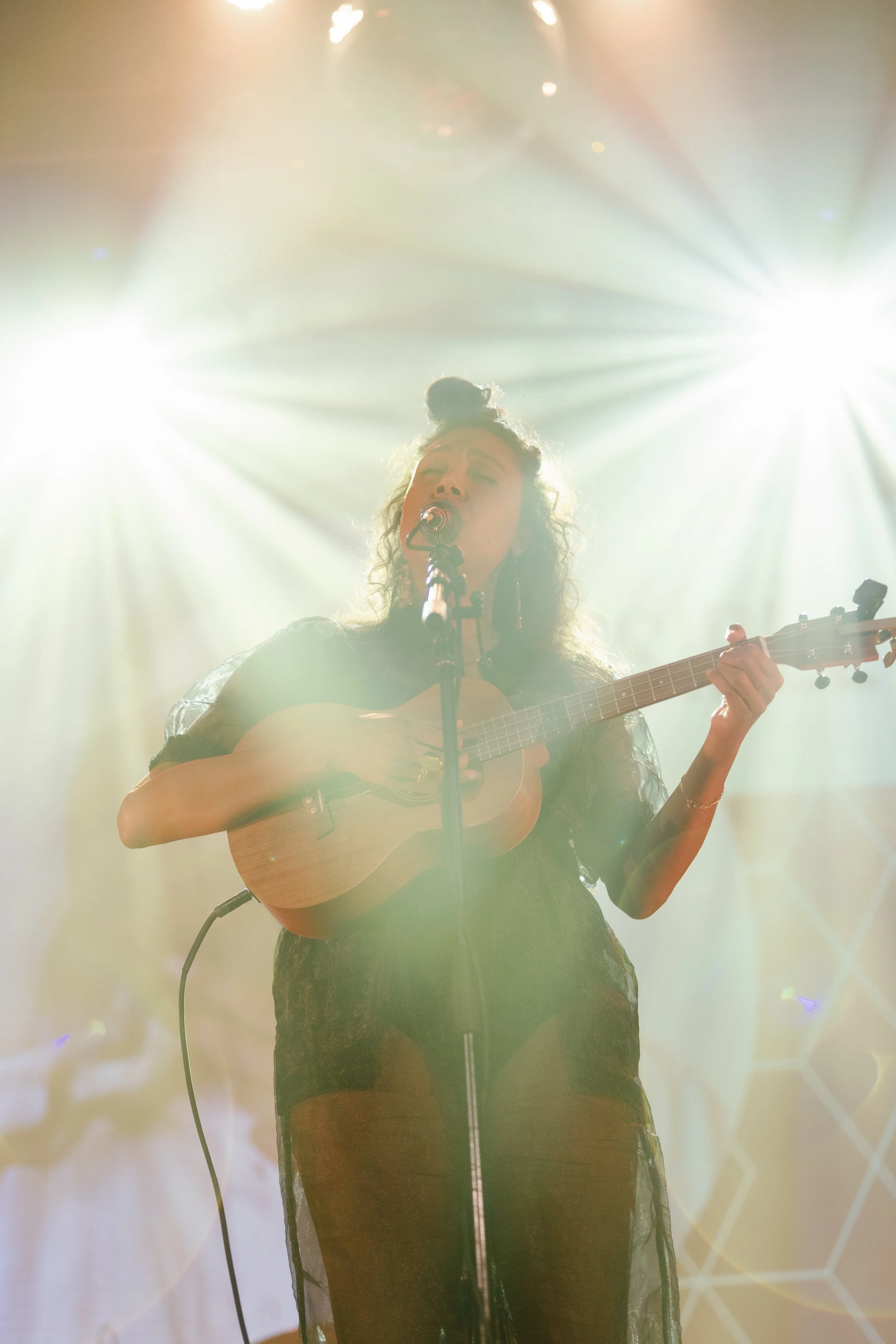 Desirée Dawson singing and playing a ukulele on stage, illuminated by bright stage lights from behind, with a patterned backdrop.