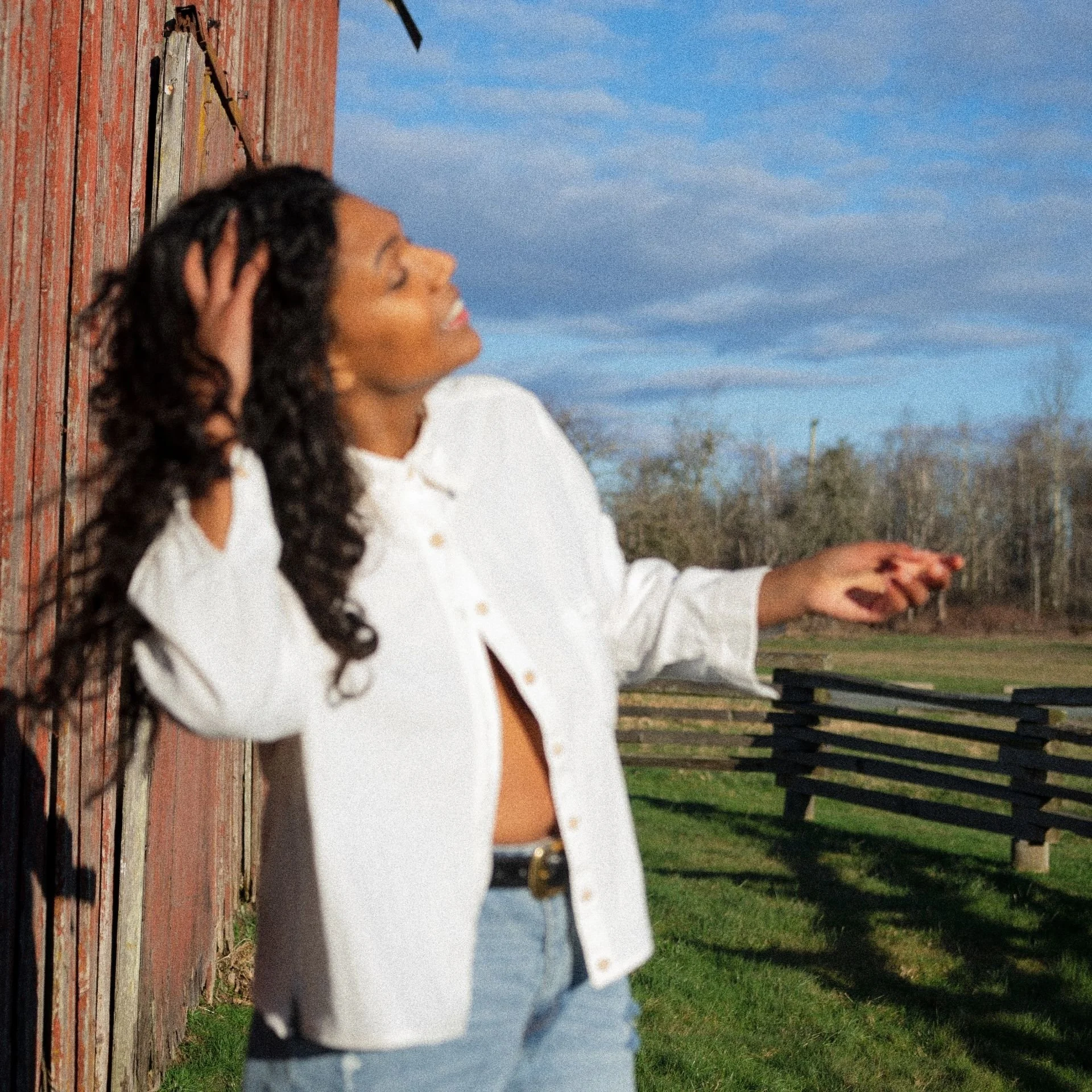 Desirée Dawson with curly black hair standing outdoors near a red wooden building, with her head tilted back, eyes closed, and one hand in her hair. She is wearing a white button-up shirt and light blue jeans.
