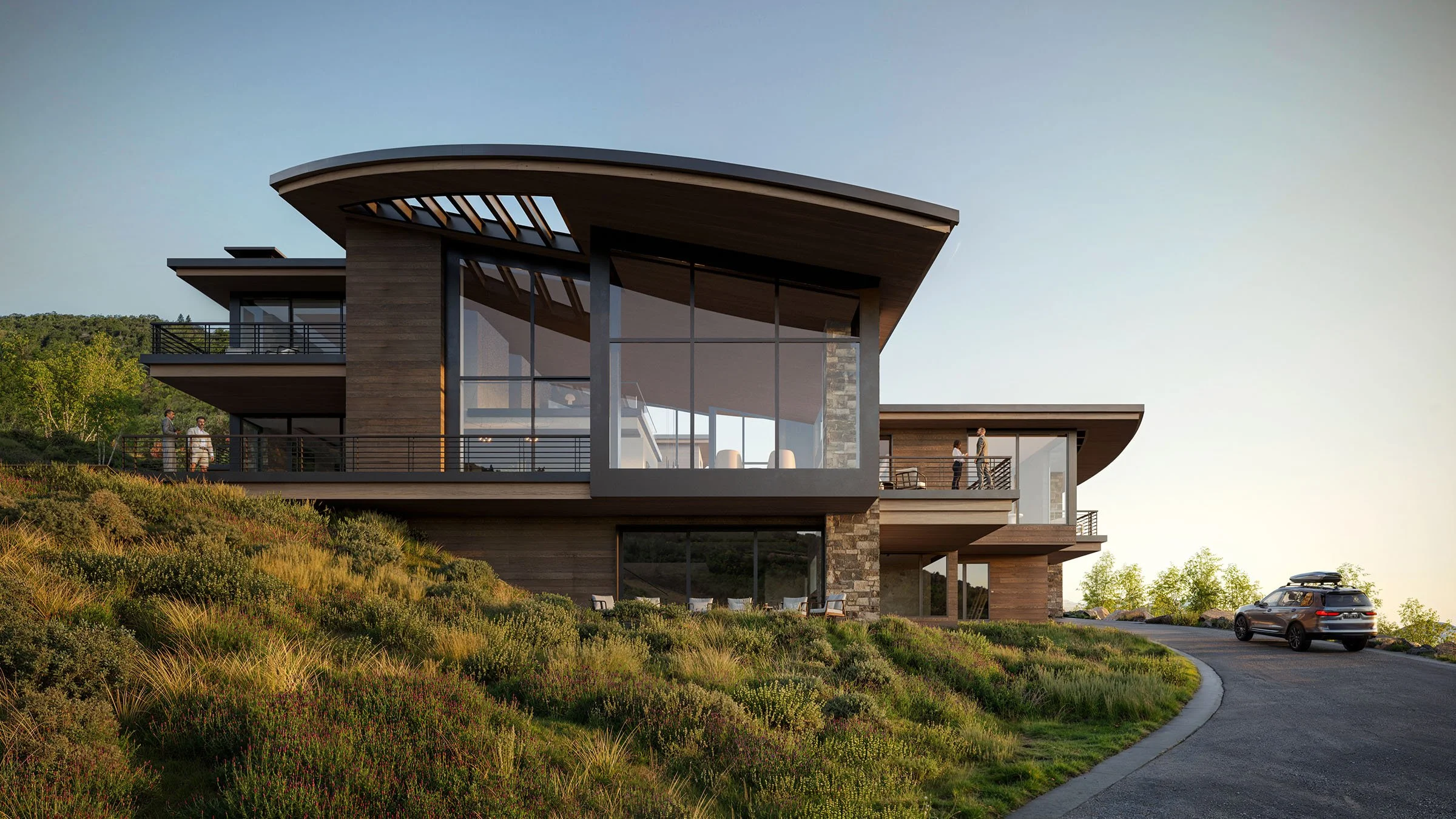 Modern house with large glass windows on a hillside, surrounded by landscaping and a curved driveway, with three people on the balcony and a car parked outside.