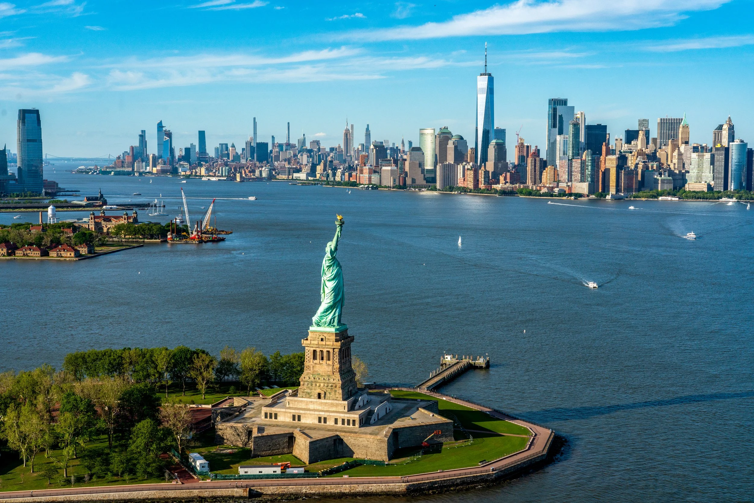 Liberty in the Harbor
A high-angle perspective of the Statue of Liberty standing guard in New York Harbor. The shot places Lady Liberty in the context of the vast waterway, with the distant shores of New Jersey and the industrial activity of the port