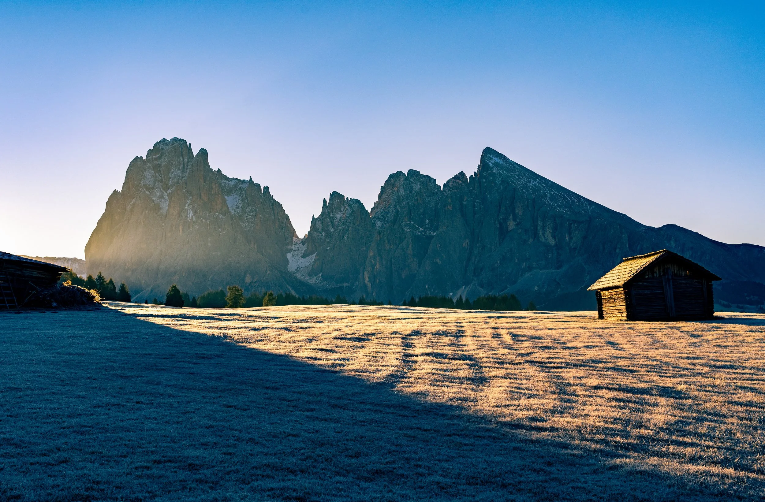 Alpe di Siusi Glow
A breathtaking view of the Alpe di Siusi at sunrise. The low-angled light rakes across the frost-covered meadow, highlighting the rolling terrain and a solitary mountain hut. In the background, the jagged peaks of the Sassolungo gr