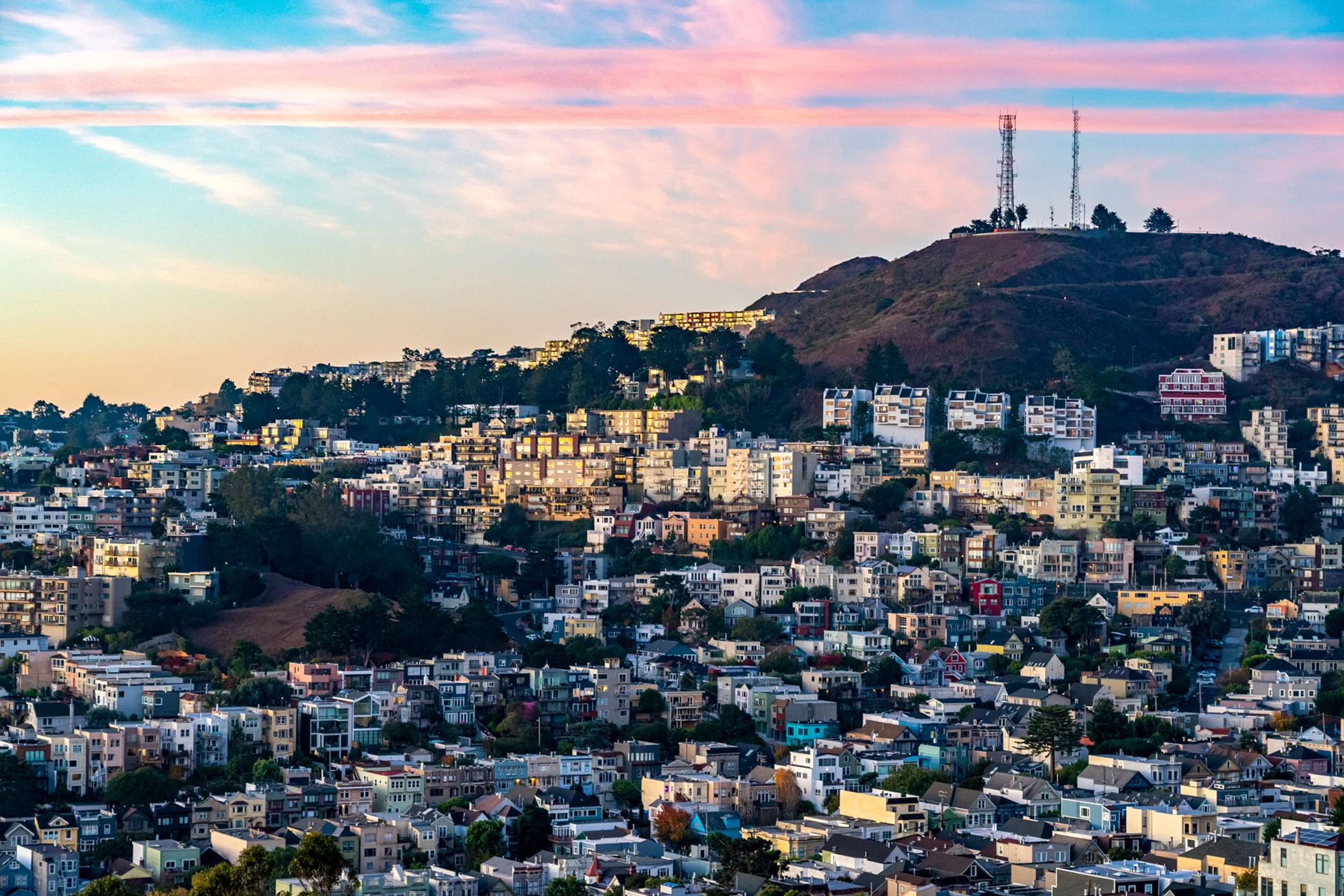 Sunset Over Corona Heights
A beautiful sunset view looking toward Corona Heights Park. The low sun casts a warm, pink-hued light over the dense residential neighborhoods, highlighting the city’s famous rolling hills and eclectic architecture. The rad