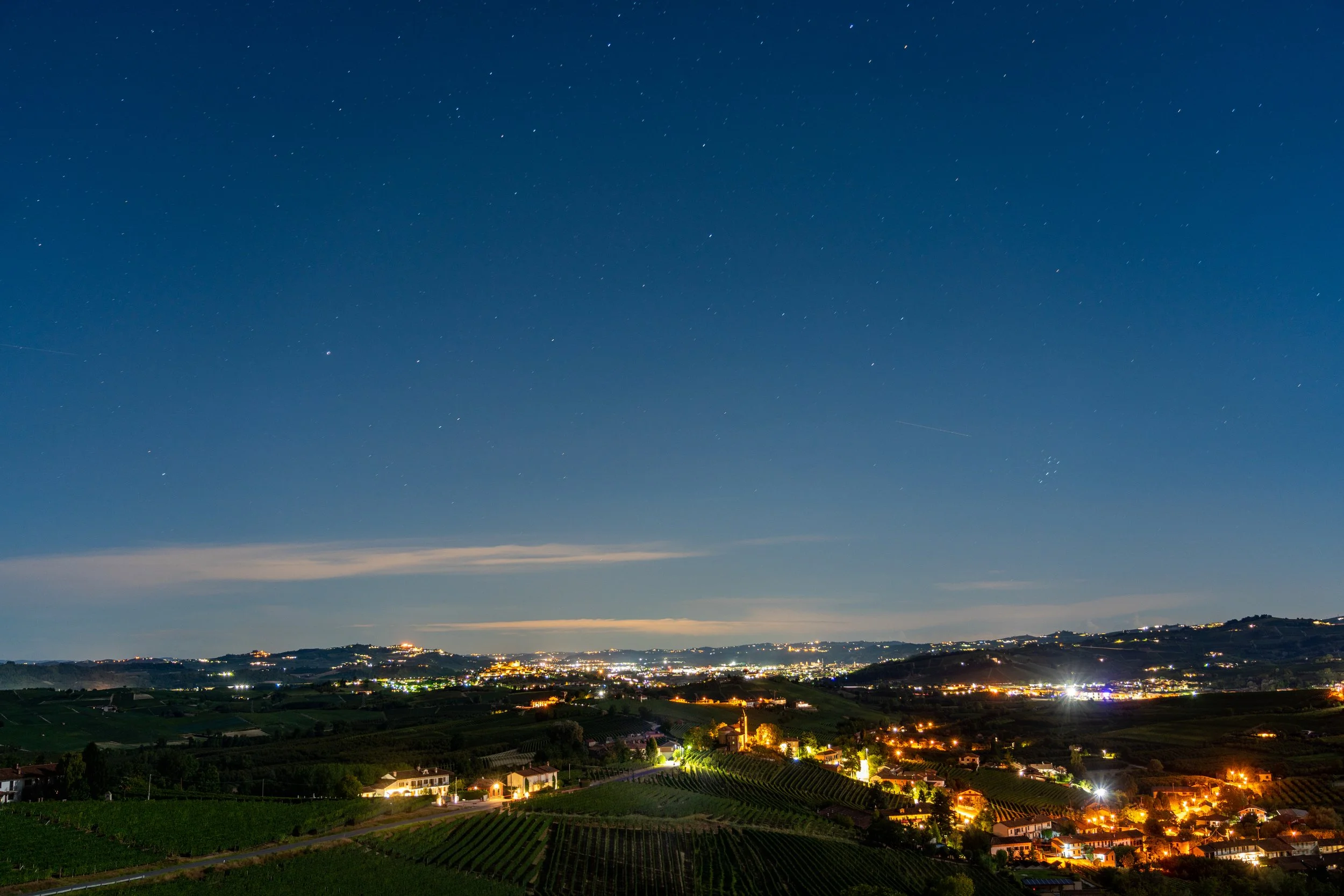 Piedmont Twilight
A serene view of the Piedmont region in Northern Italy as the last light of dusk fades into a deep blue night. The rolling vineyards are punctuated by the warm, amber glow of small towns and villages nestled in the hills. This shot 