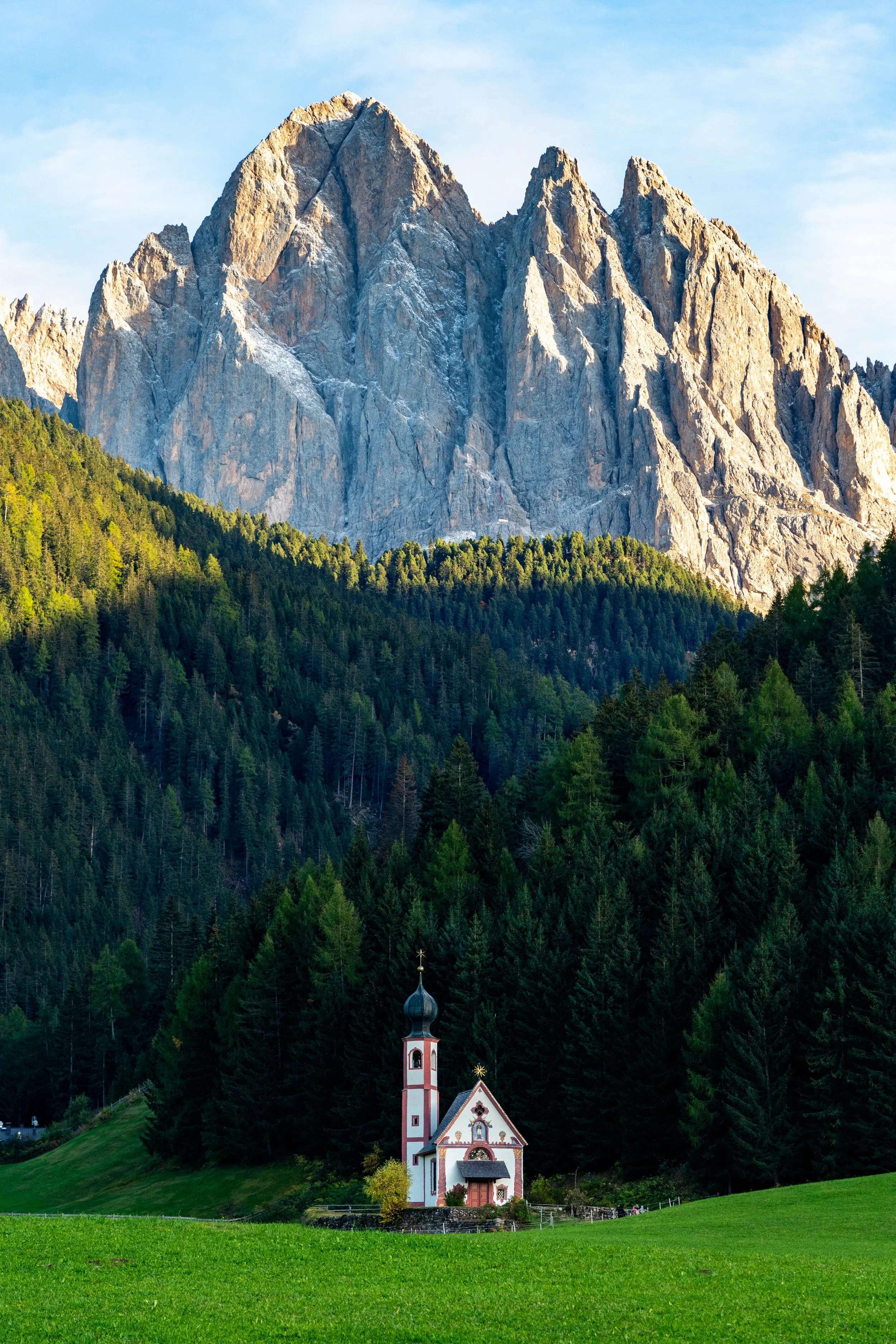 The Mountain Chapel
A picturesque view of the Church of St. John in Ranui, nestled in a lush green meadow at the base of the Odle Group mountains. The solitary Baroque chapel, framed by dark evergreen forests and the towering, jagged limestone peaks 