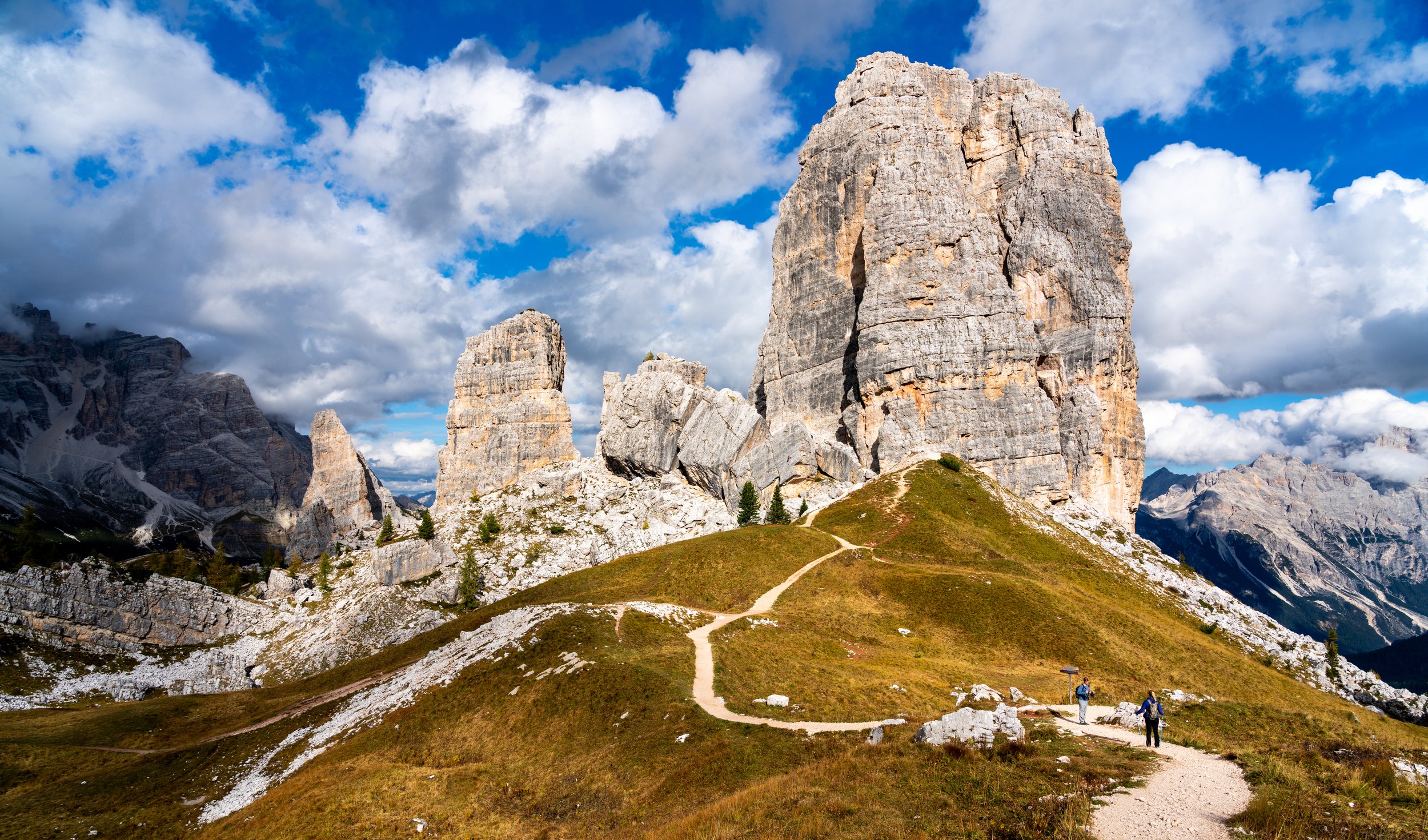 The Giants' Path
A wide-angle perspective of the winding trail leading toward the base of the Cinque Torri rock formations. The path provides a sense of scale against the massive limestone "Five Towers," while the bright, high-altitude sun brings out