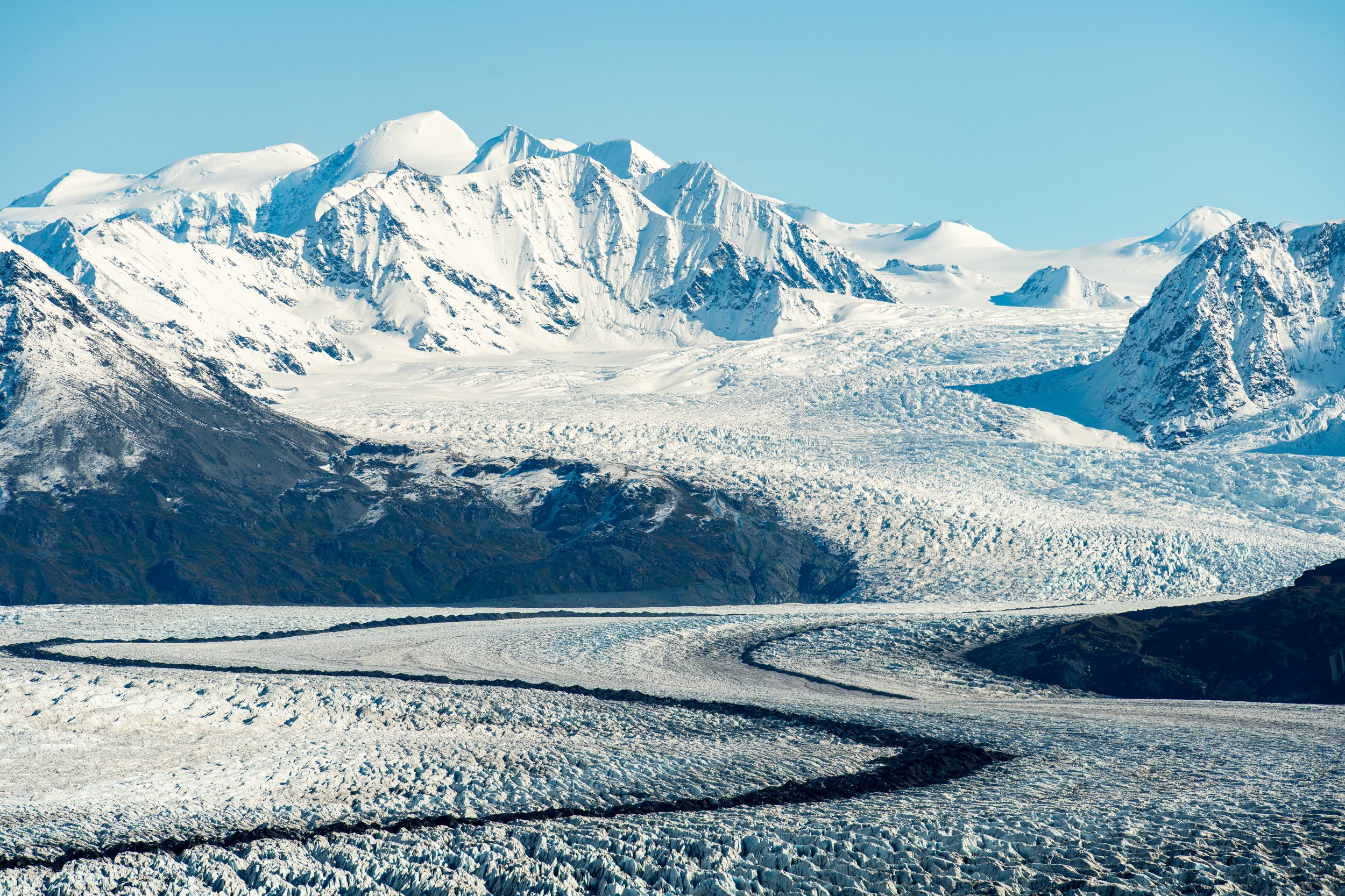 The Frozen Artery
A high-altitude view looking down at a massive glacier as it winds through a mountain pass. This shot highlights the "medial moraines"—the dark stripes of rock and soil trapped in the ice—which reveal the glacier's flow history like