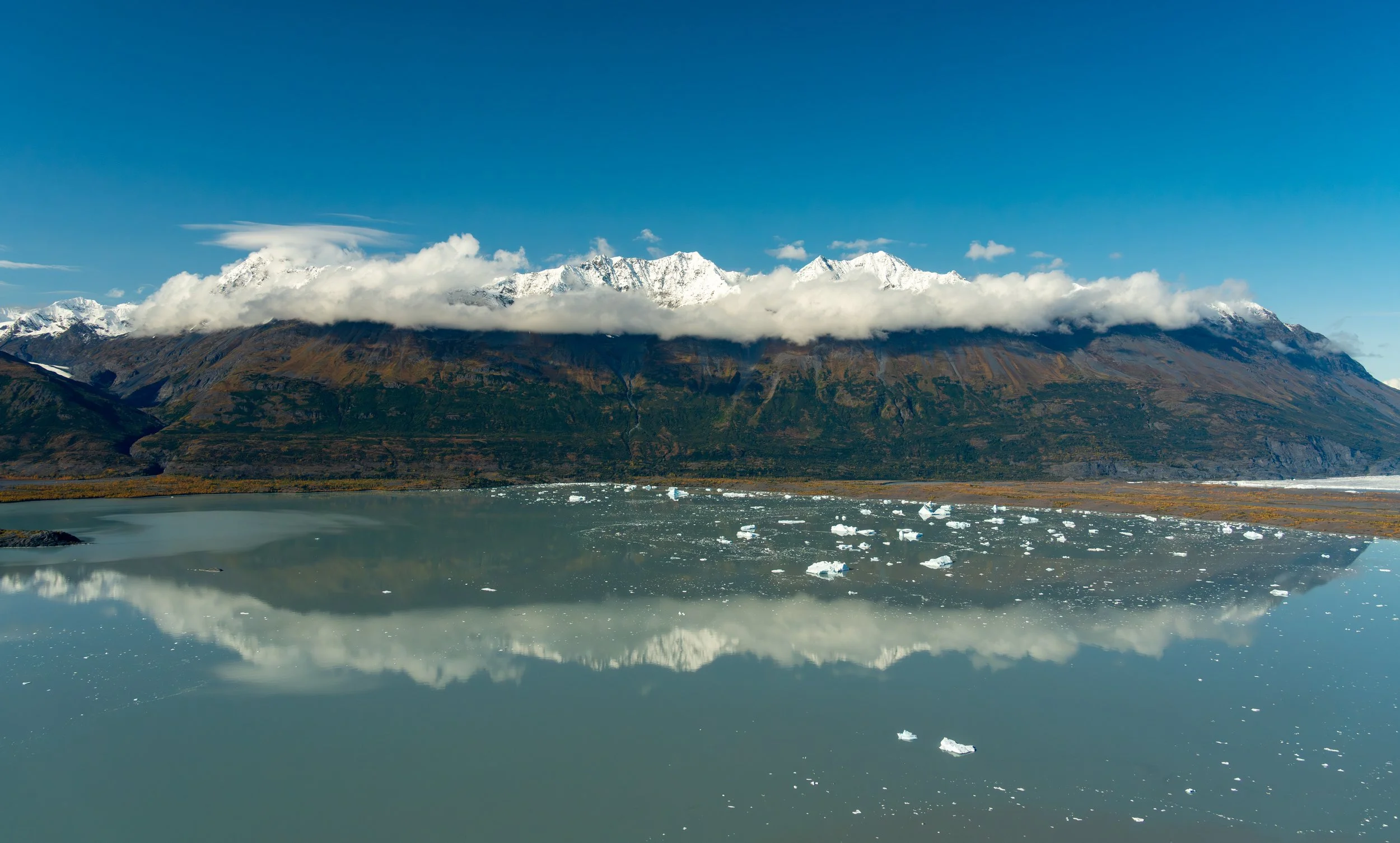Alpine Reflection
 A serene, wide-angle shot of Prince William Sound, where the rugged Chugach Mountains reflect perfectly in the calm, glacial waters. This perspective highlights the floating "bergy bits" that have calved from nearby glaciers, empha