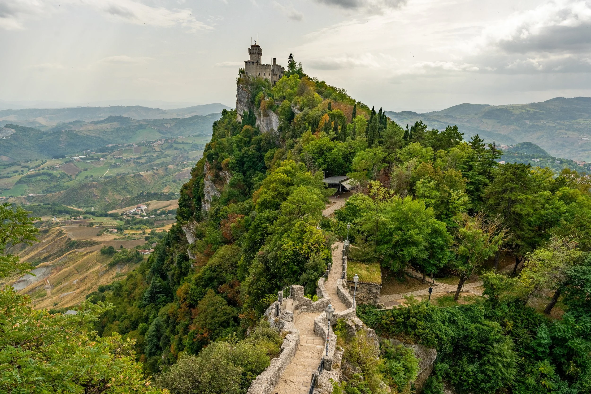 The Citadel
A view of the Cesta Tower (the Second Tower) perched on the highest peak of Mount Titano. The shot captures the ancient stone fortifications as they follow the natural, jagged curve of the cliffside, with the rolling green valleys of San 