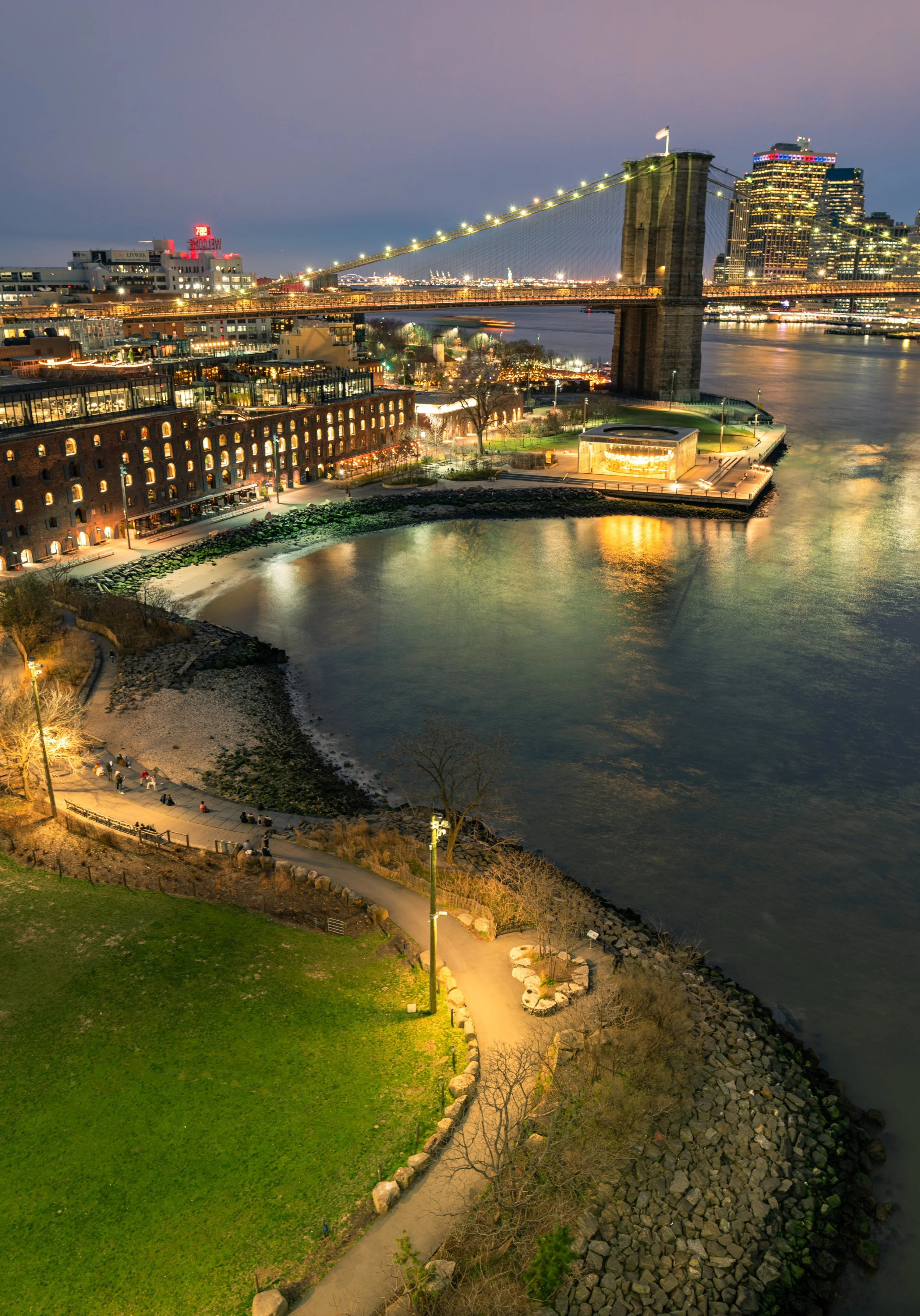The Cobblestone Curve
A high-angle view looking down at the intersection of the DUMBO waterfront and the East River. The shot captures the warm glow of the streetlights reflecting off the cobblestone streets and the quiet green spaces of Brooklyn Bri