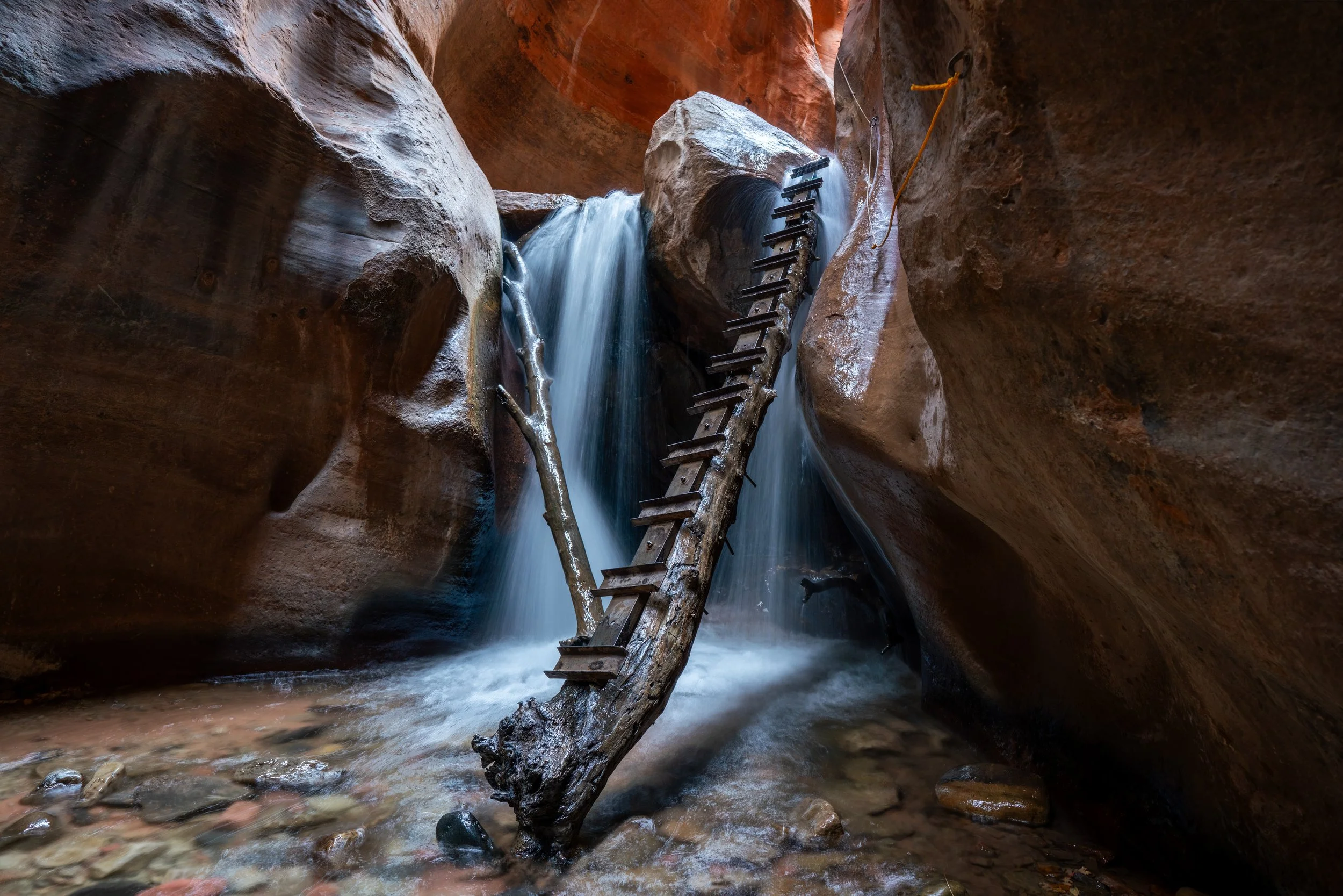 The Hanging Log
Deep within the narrow slot of Kanarra Creek, a solitary waterfall cascades over an ancient, wedged log. The vibrant orange of the sandstone walls, carved by centuries of water, contrasts sharply with the cool, rushing flow of the fal