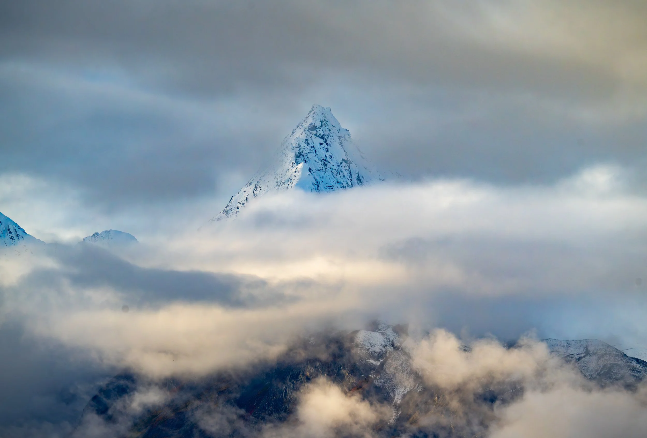 Roof of North America
A stunning perspective of the Denali mountain range, captured as the summit pierces through a layer of soft, atmospheric clouds. This high-altitude shot emphasizes the sheer scale and icy isolation of the Alaska Range, where the