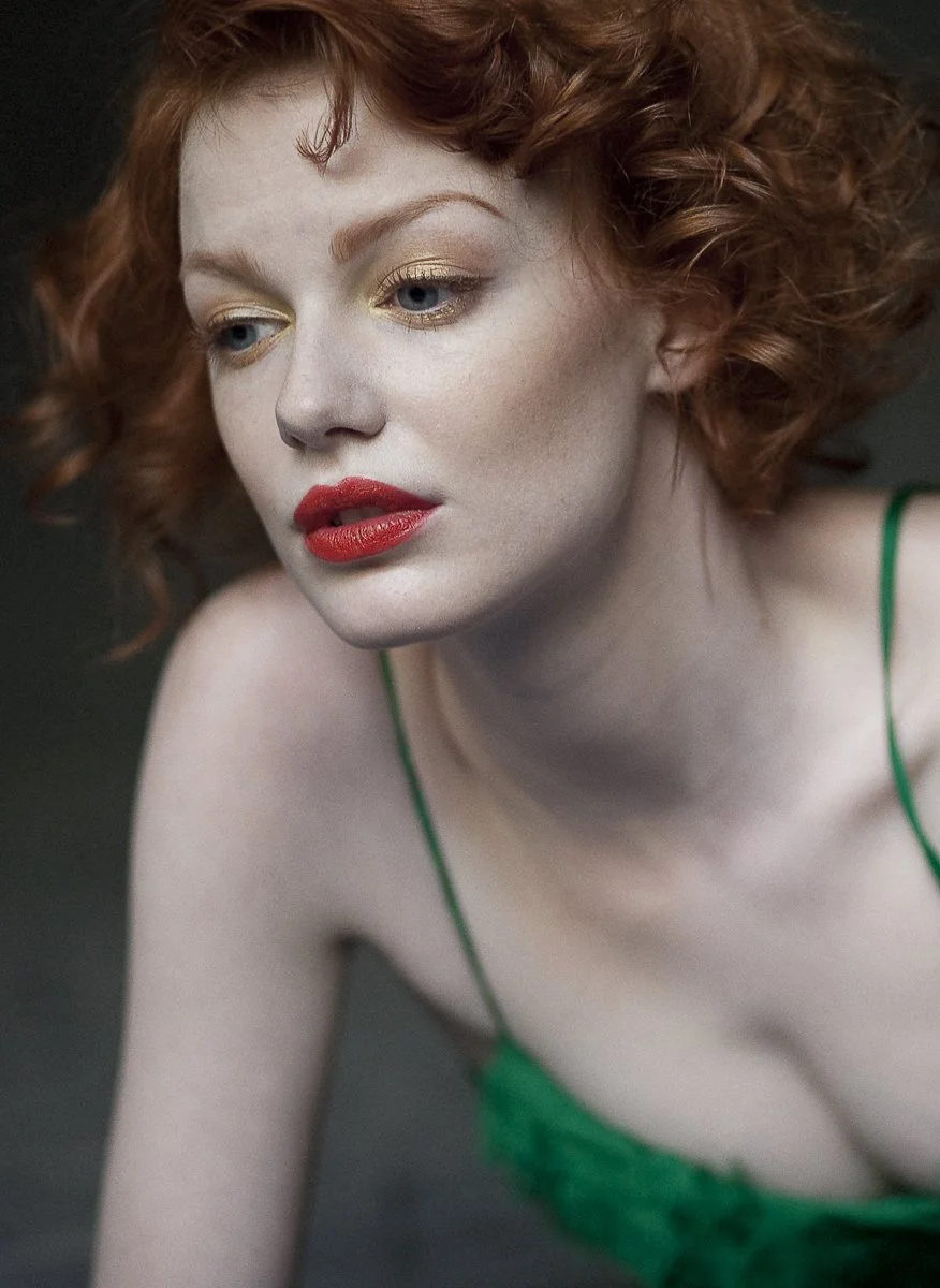A woman with curly red hair and fair skin wearing a green spaghetti strap top, with makeup including red lipstick and gold eyeshadow, looking off to the side.