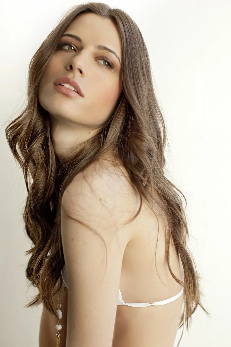 A young woman with long, wavy brown hair looking sideways against a plain, light background.