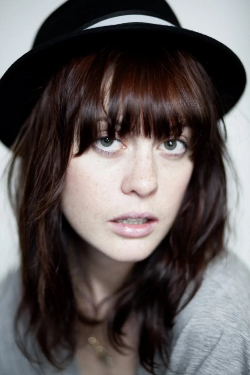 Close-up of a young woman with red hair, wearing a black hat, looking at the camera with a neutral expression.
