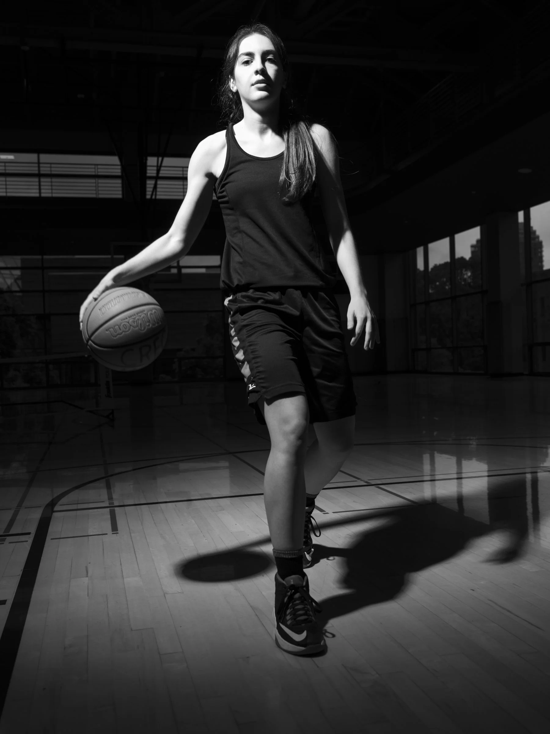 A young woman wearing a sports tank top, shorts, and high-top sneakers is dribbling a basketball on a gymnasium basketball court. The image is in black and white, with high contrast lighting highlighting her and casting shadows.