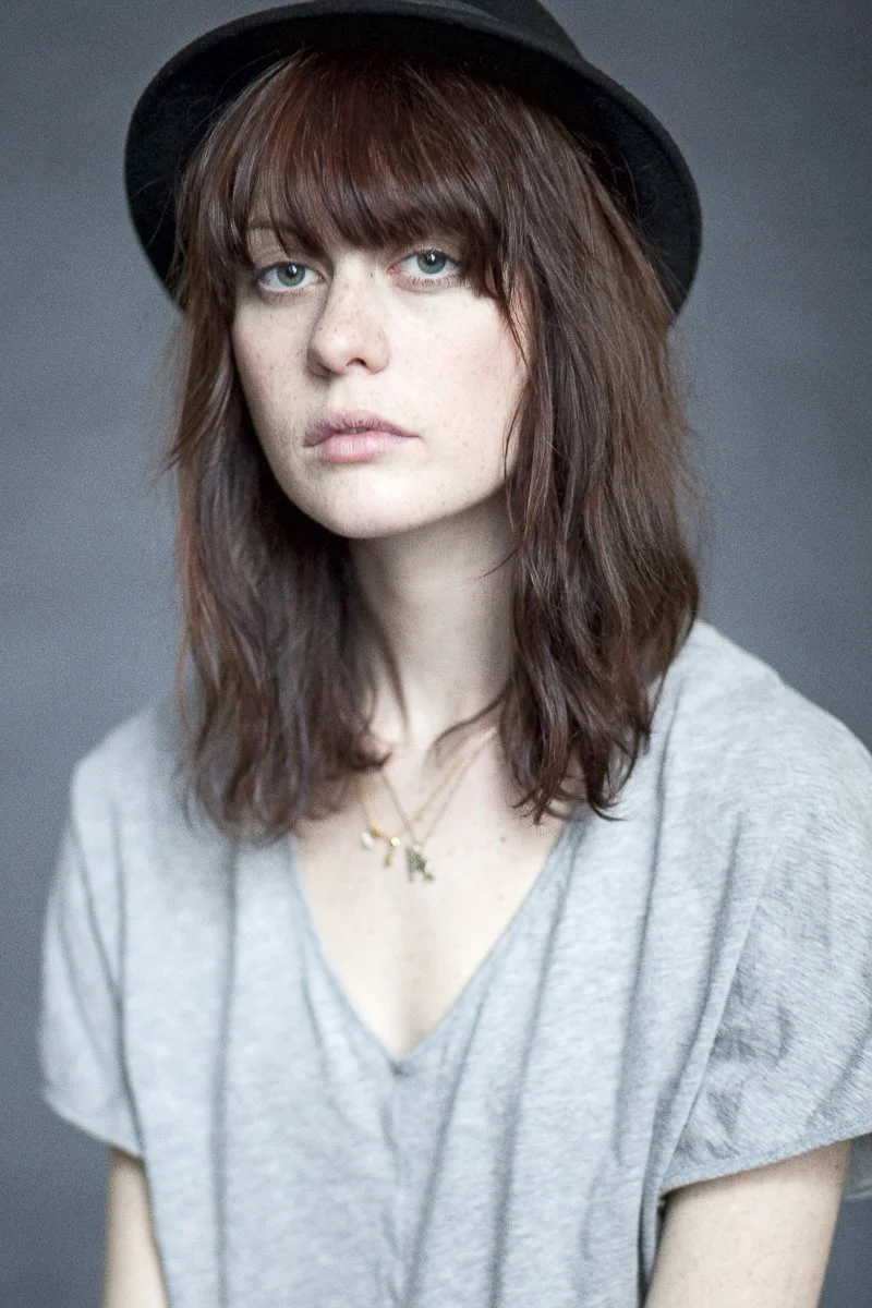 A young woman with blue eyes, reddish-brown wavy hair, wearing a black hat and a light gray t-shirt against a dark gray background.
