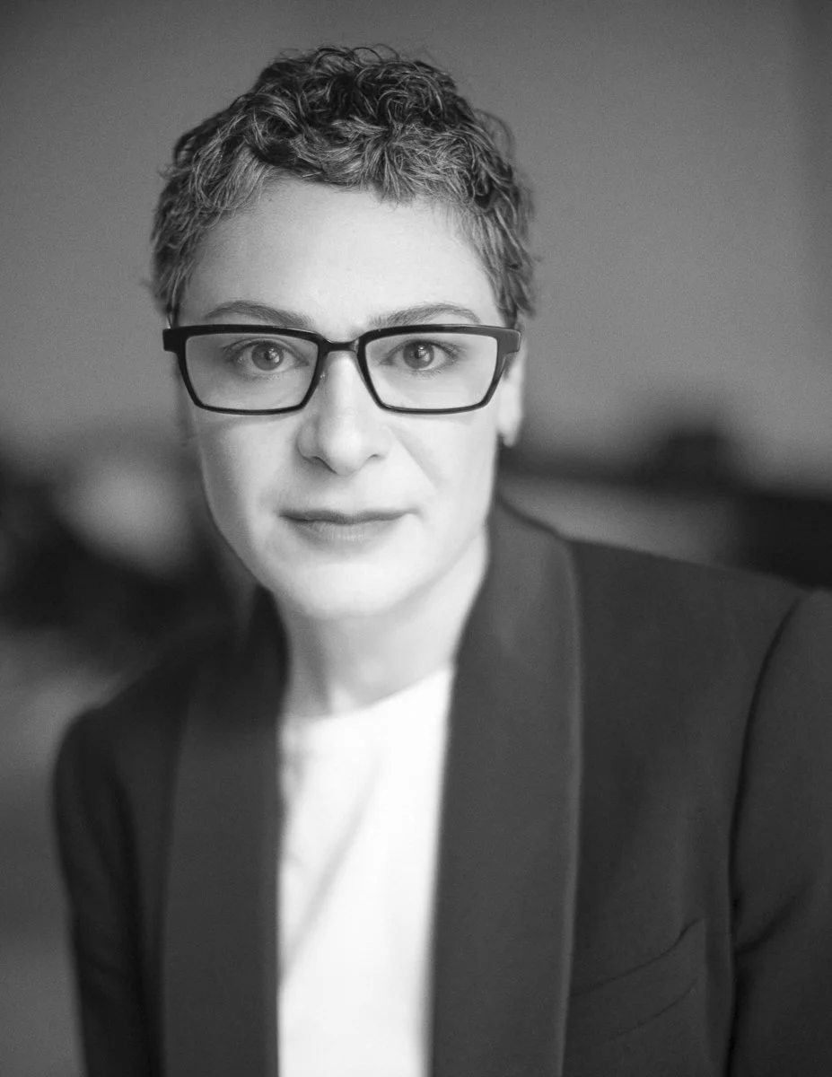 Black and white portrait of a woman with short curly hair, wearing glasses and a blazer, looking at the camera.