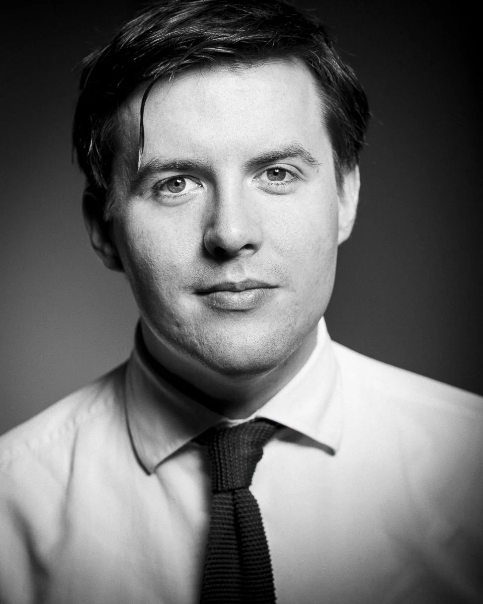 Black and white portrait of a young man with medium-length dark hair, wearing a white shirt and a dark tie, looking directly at the camera with a neutral expression.
