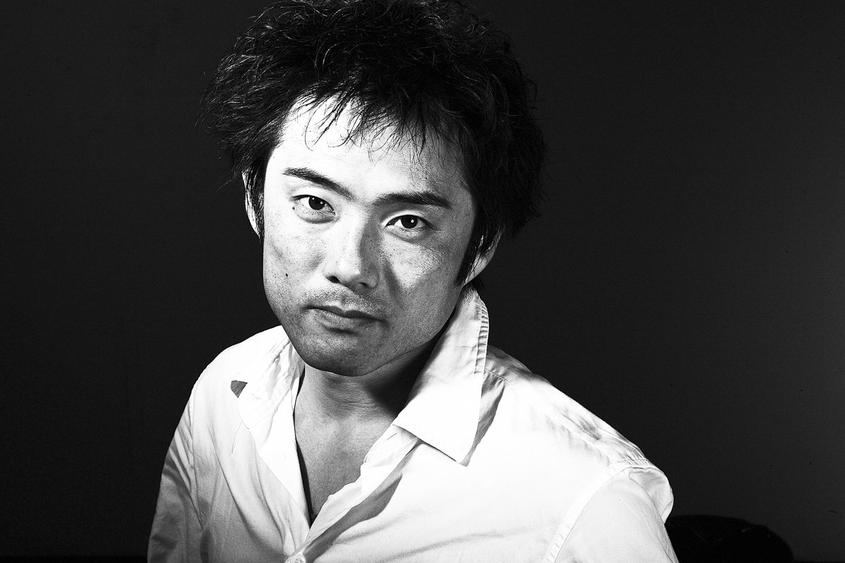 A black and white portrait of a man with messy hair, wearing a white shirt, against a dark background.